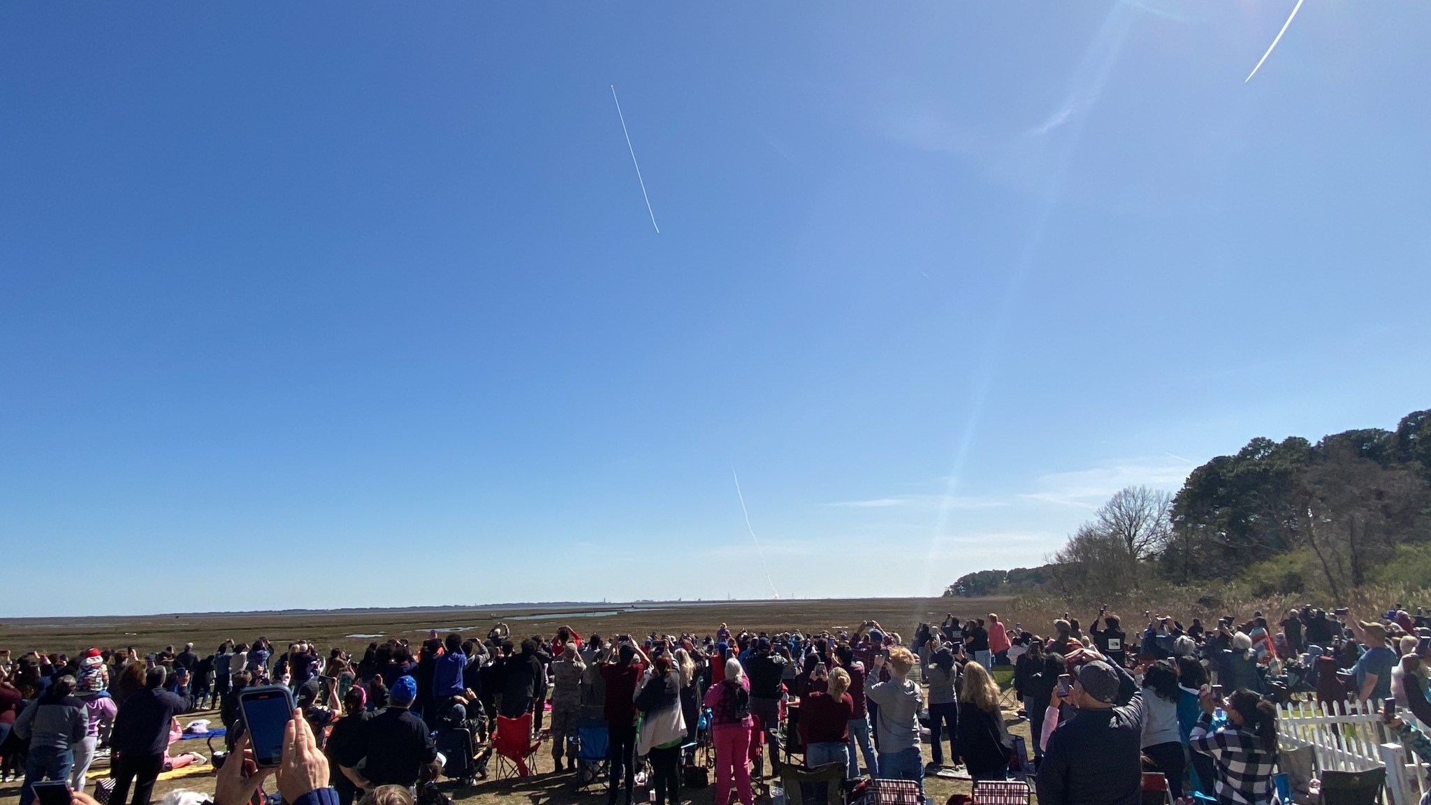 A group of people stand facing away from the camera some holding cell phone cameras above their heads watching a rocket launch from Wallops Island