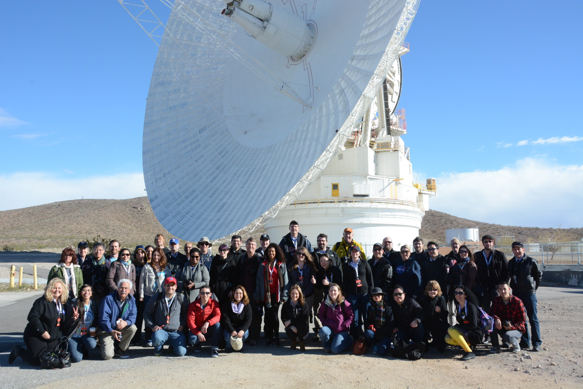 A group of people pose beneath a Deep Space Network antenna at the Goldstone Deep Space Communications Complex.