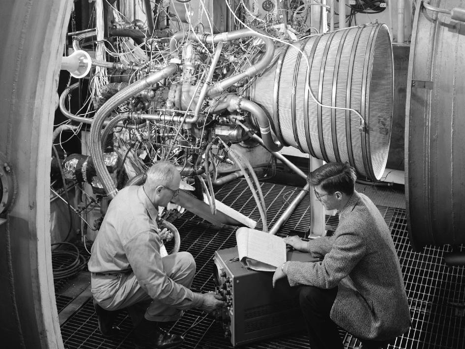Two men squatting next to rocket engine.
