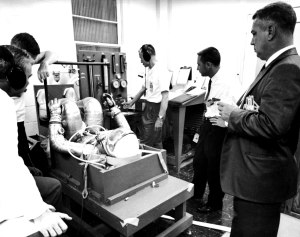 Alan Shepard, suited in his silver space suit, sits horizontally in flight couch as 5 men help prepare him for his flight.