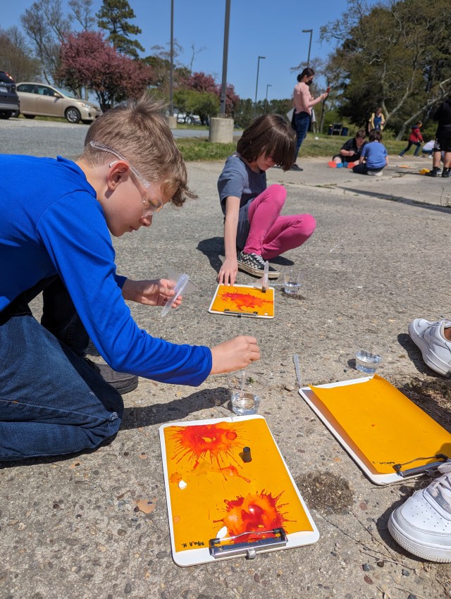 A fourth grade boy wearing clear safety goggles, a blue long sleeve shirt and dark jeans, kneels over a piece of goldenrod paper splattered with bright red patches from water reacting with the paper. The student is carefully placing calcium carbonate tables into a test tube for a rocketry experiment