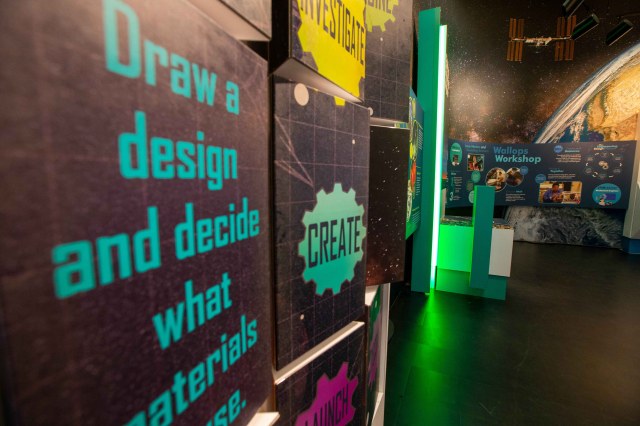An engineering exhibit featuring bright turquoise writing on a black background, only part of the sentance is visible, it says draw a design and decide what materials to use