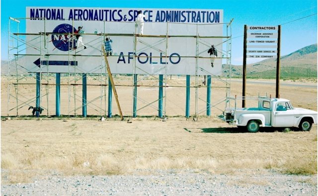 Workers use scaffolding to install a billboard that says National Aeronautics and Space Administration and Apollo with an arrow pointing to the left.