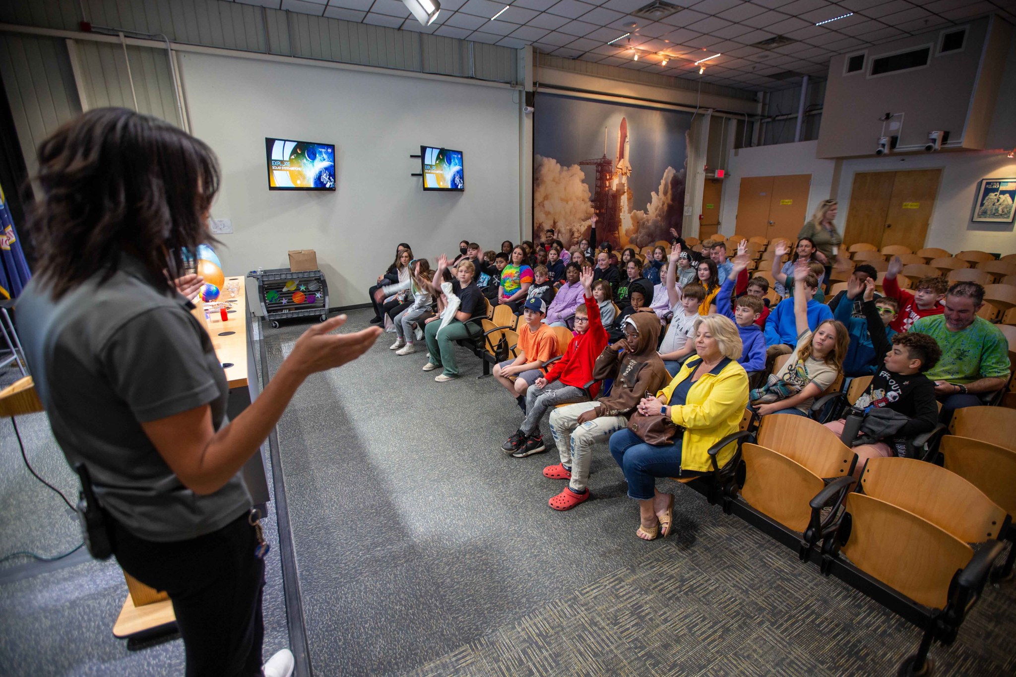 A educator stands in front of a group of students who are raising their hands to answer a question