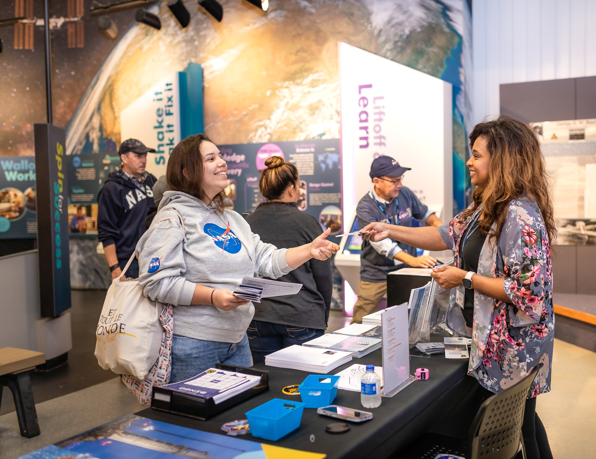 A woman standing behind a table is handing an informational brochure to a guest at a museum