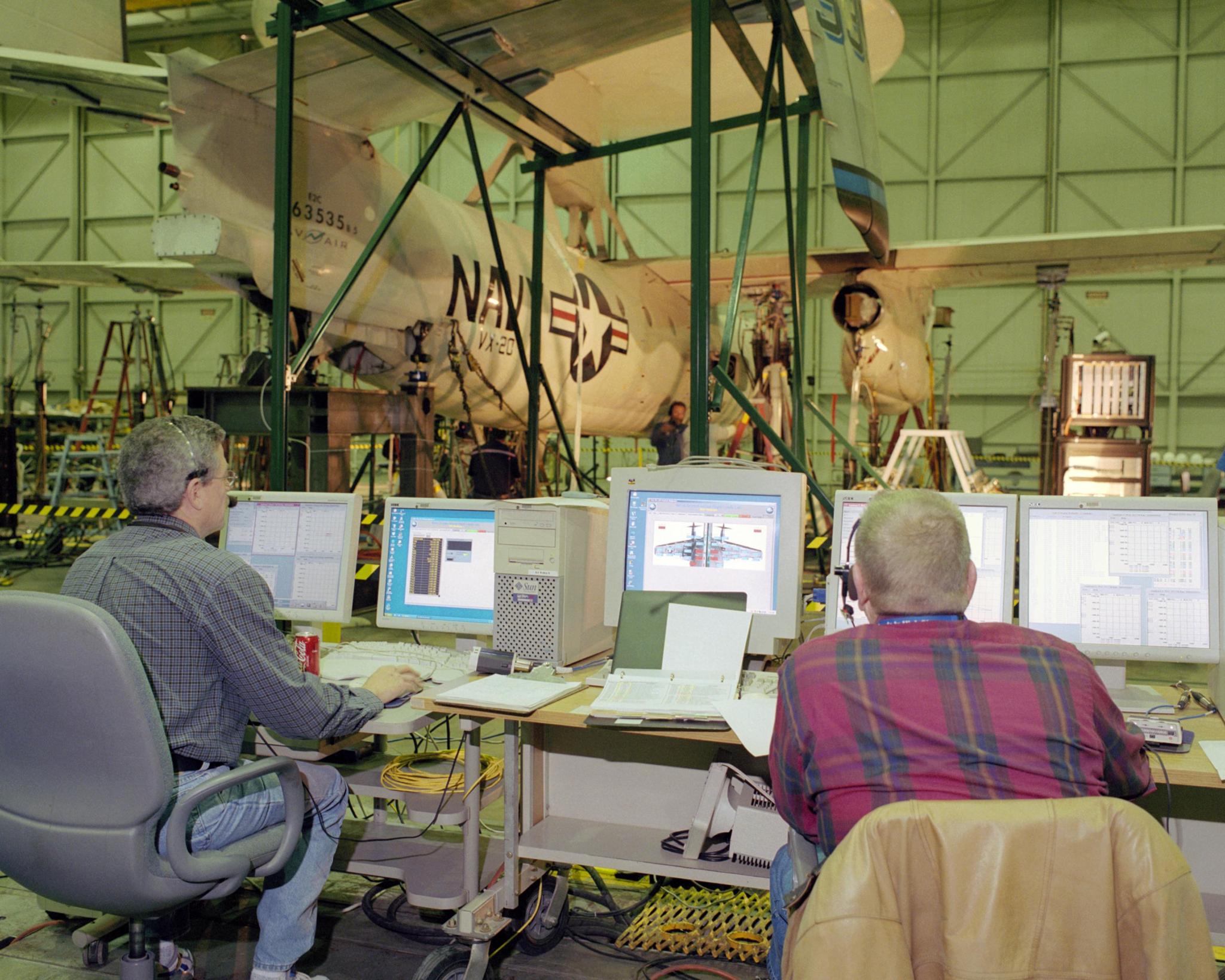 Two men sit at computers while testing an airplane in the background