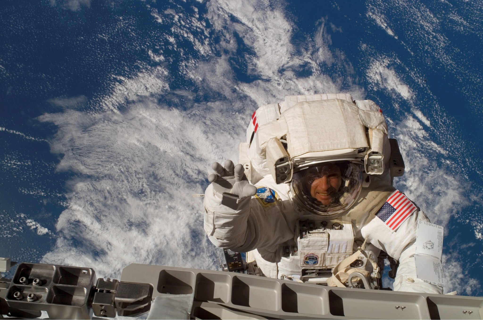 An astronaut dressed in a large, bulky spacesuit waves to the camera while working in space