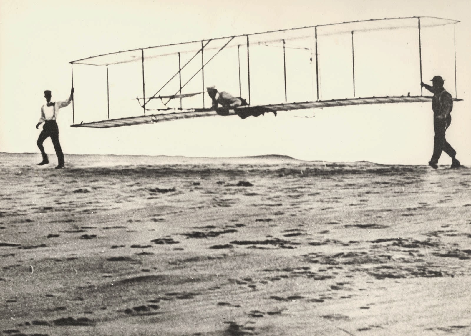 Historic photo of the Wrights’ third test glider being launched, with one man aboard and one to each side of the glider