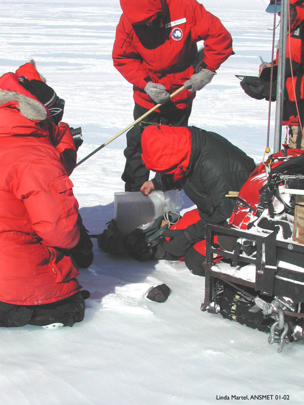 A small black meteorite lies in the snow while a man dressed in black and red winter clothing documents where it was found