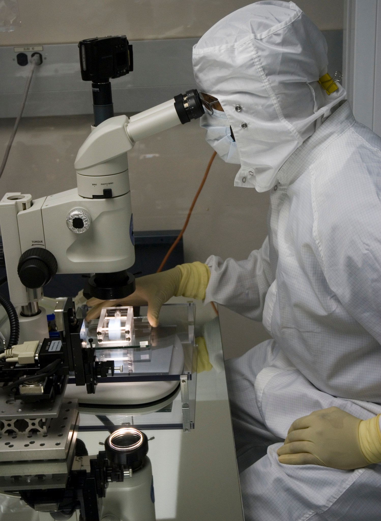A person with protective yellow gloves and white lab clothes, including mask and head covering, looks into a microscope