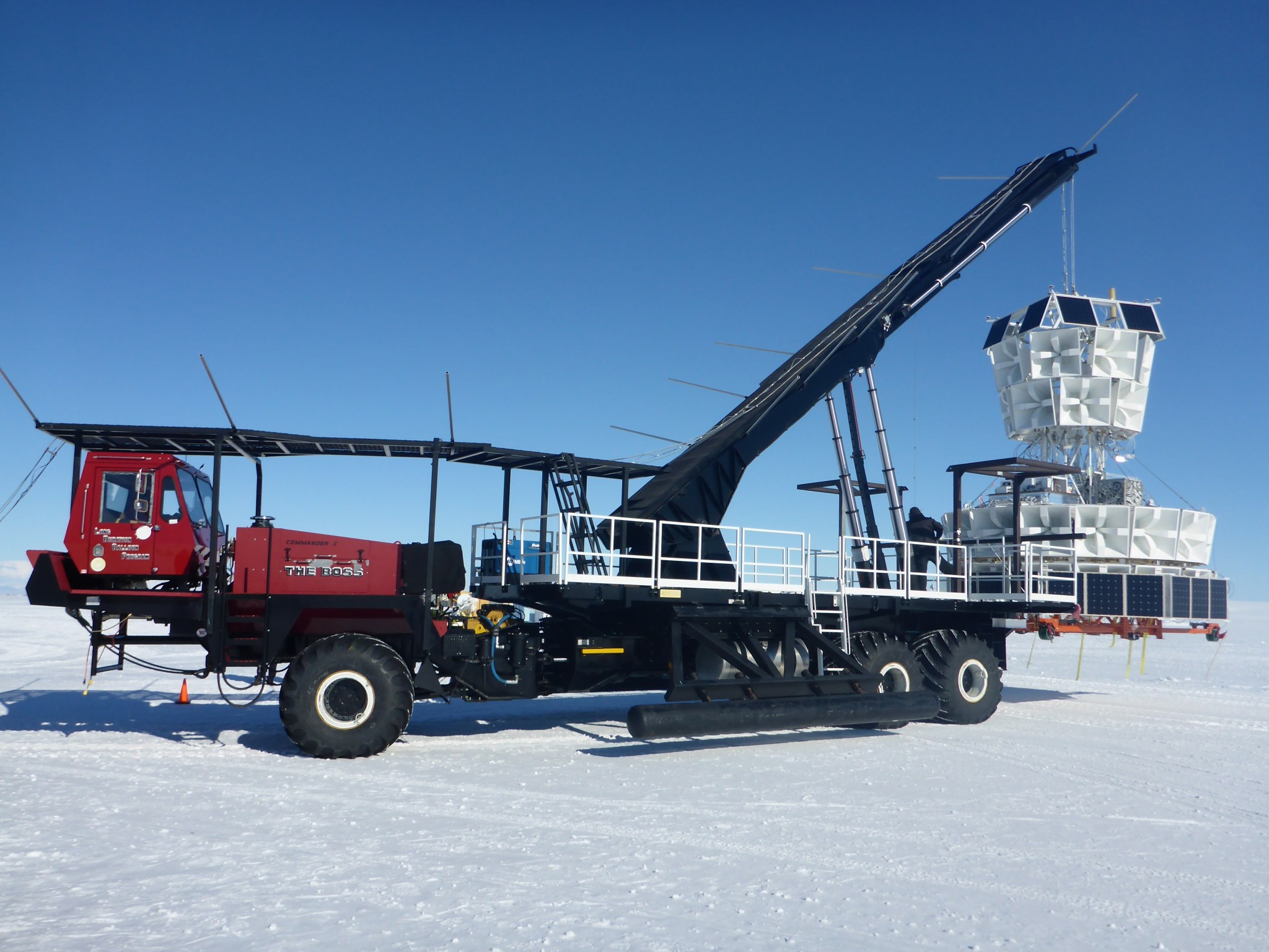 A crane holds the ANITA-III payloada on a snowy landscape. The payload is made up of mostly white indented cubes, with small solar panels ontop.