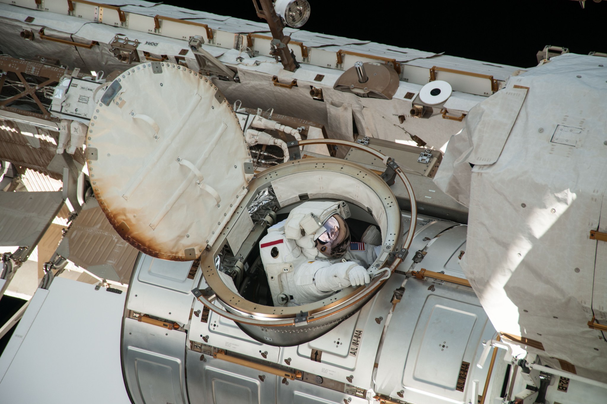 Astronaut wearing a bulky white spacesuit looks out at space from the open hatch of the space station's airlock