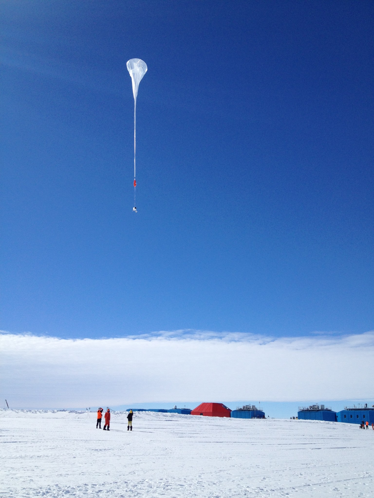 A white balloon floats into the sky as three people watch from the snow-covered ground