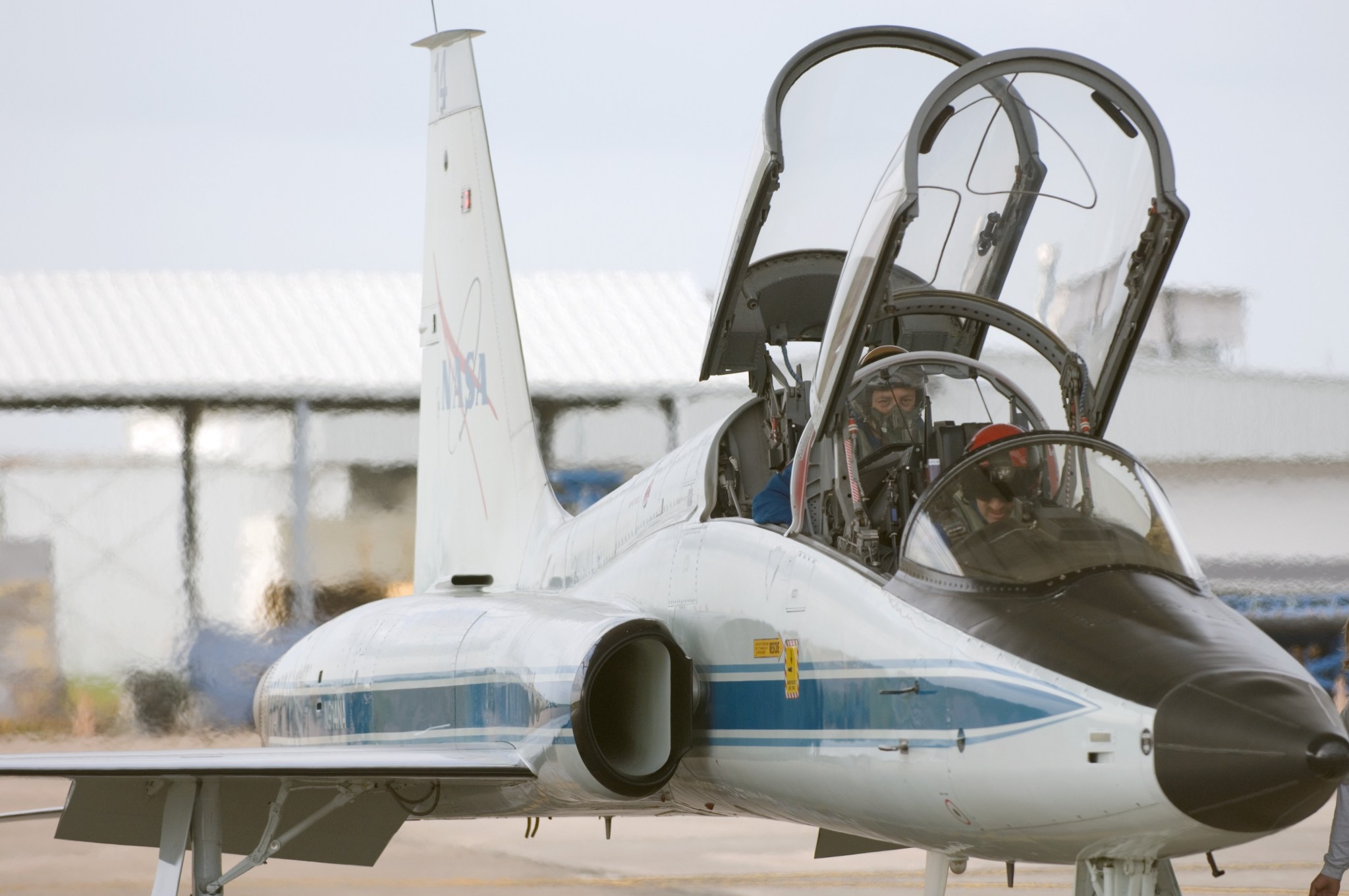 A small white jet sits on a runway with two astronauts in its seats and the two canopies opened
