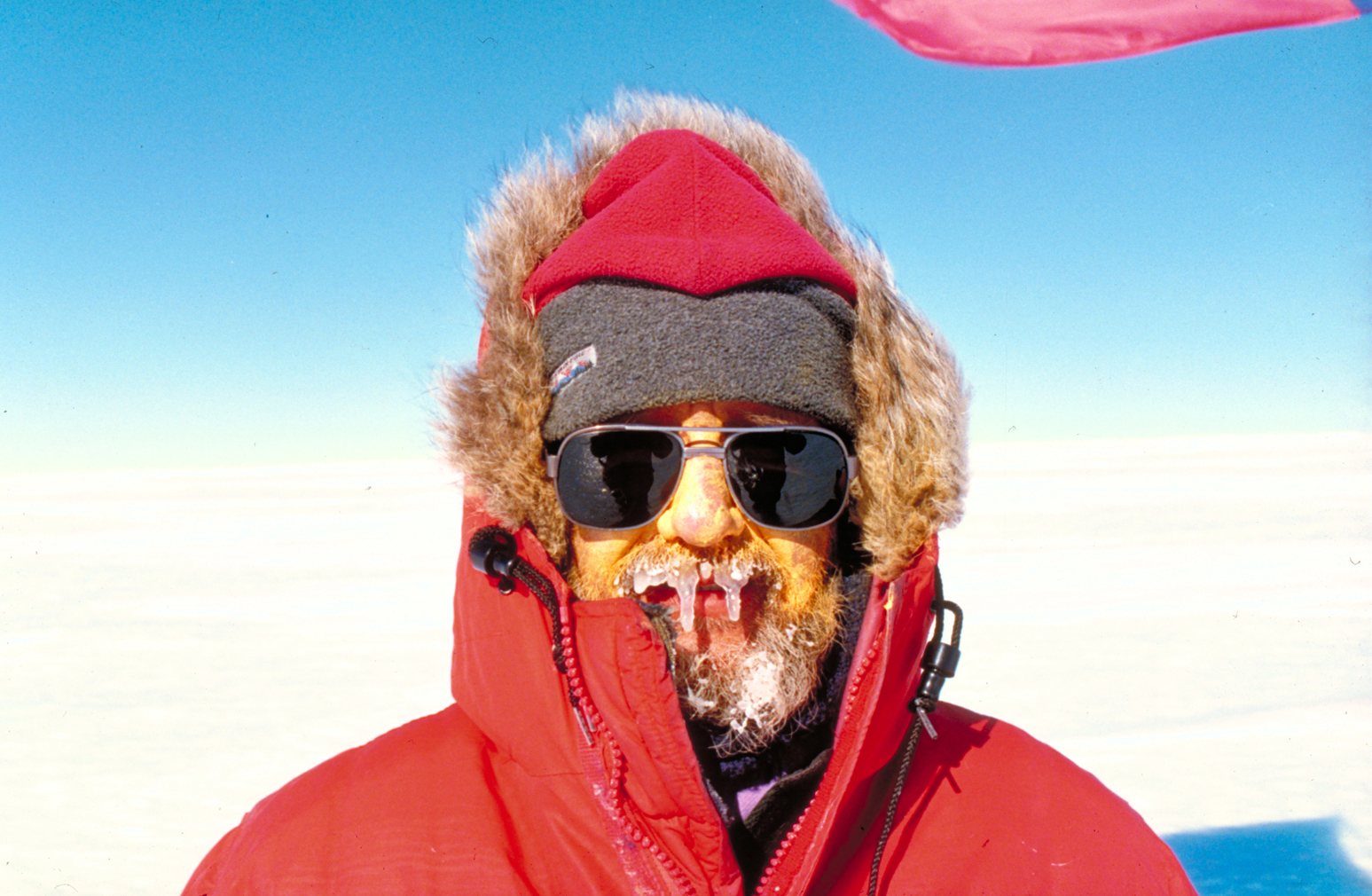 Close-up picture of a man outdoors in Antarctica dressed in heavy, red winter clothing with ice on his face
