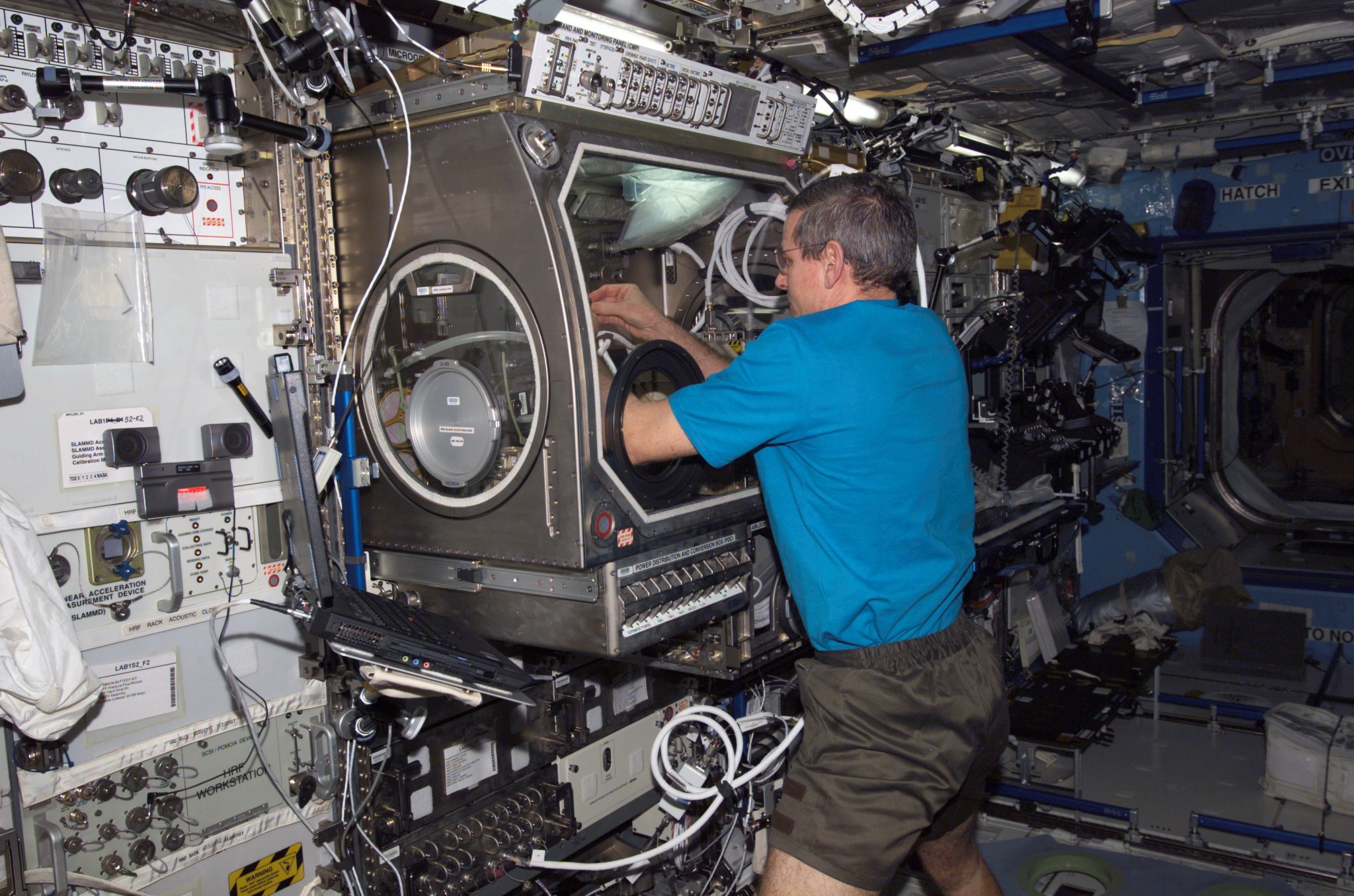 Wearing a short-sleeved shirt and shorts, an astronaut puts his hand in a glovebox