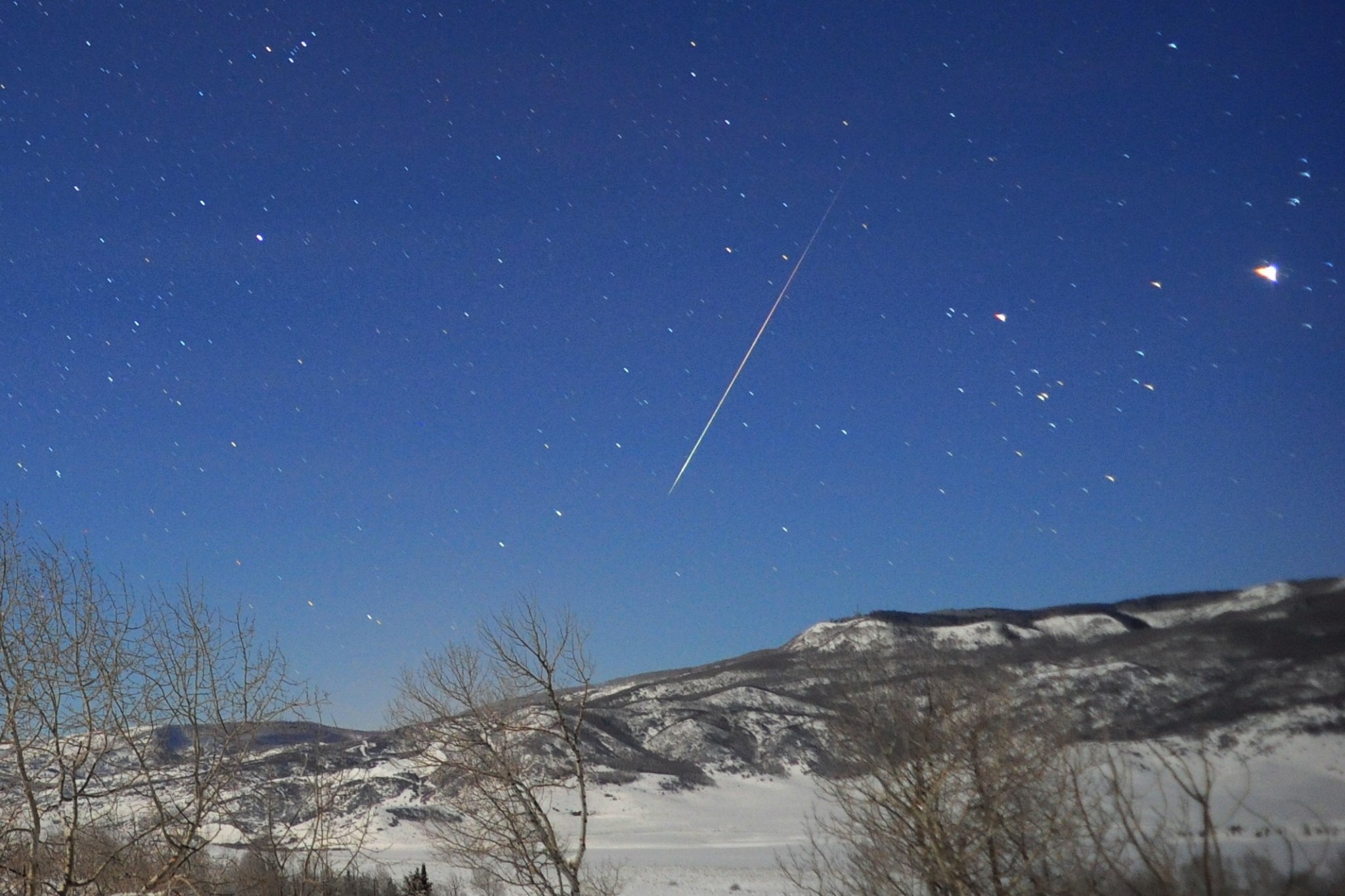 A meteor streaks across a blue sky above a rugged, snowy landscape
