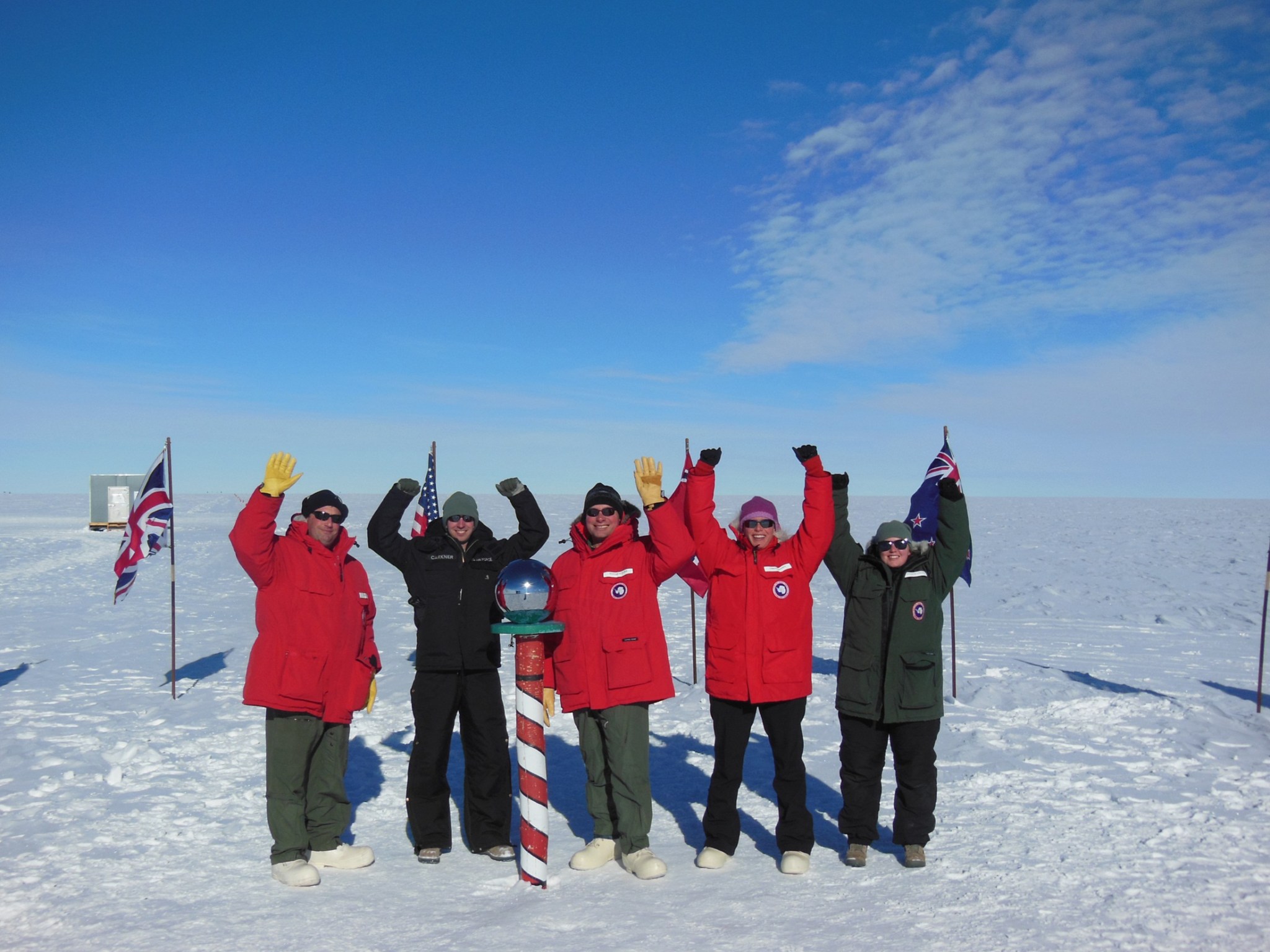 Five people dressed in cold weather gear stand outside in the snow behind a red and white pole marking the South Pole