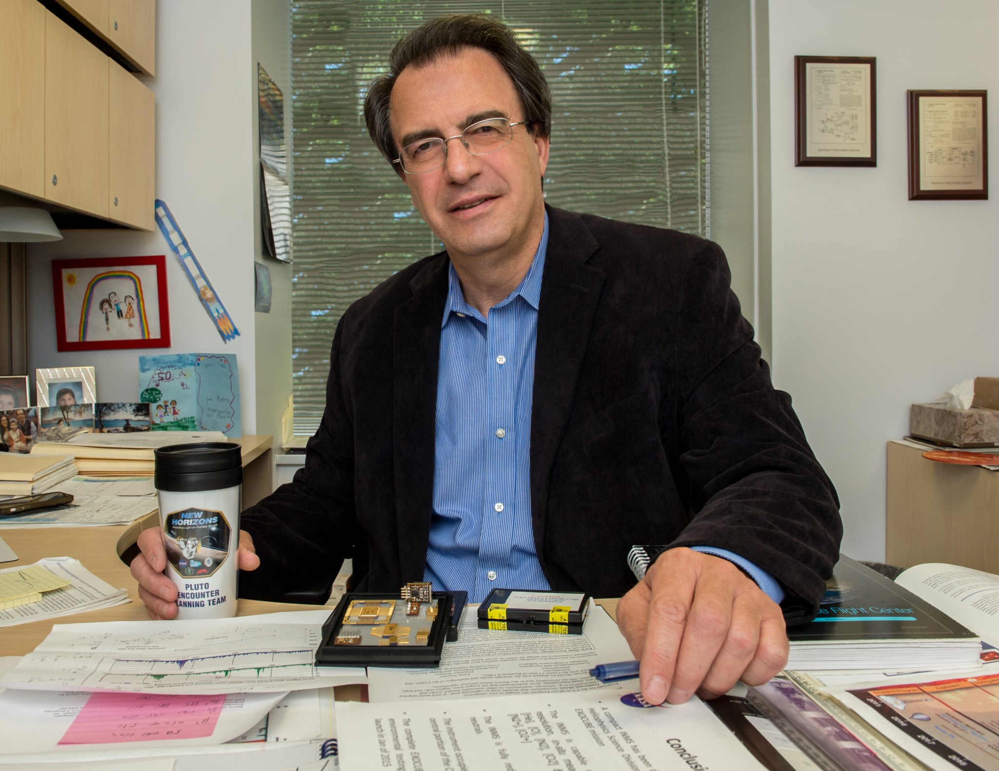 Nikolaos Paschalidis sits at a desk in an office, facing the camera. In one hand he holds a mug with the New Horizons mission logo on it.