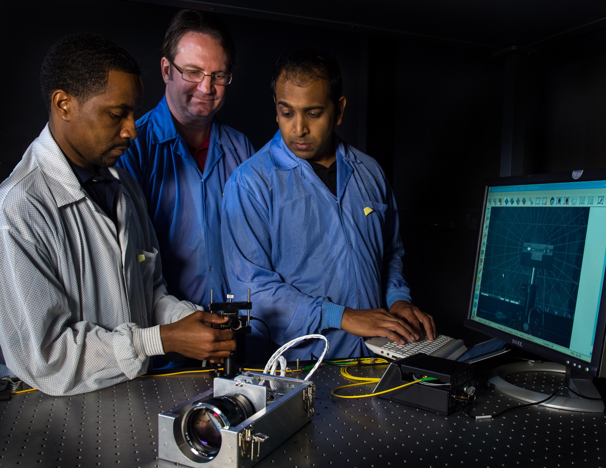 Three men in lab coats stand in front of a black table, looking at a small satellite with a big lens on it. One of them is working on a computer keyboard.