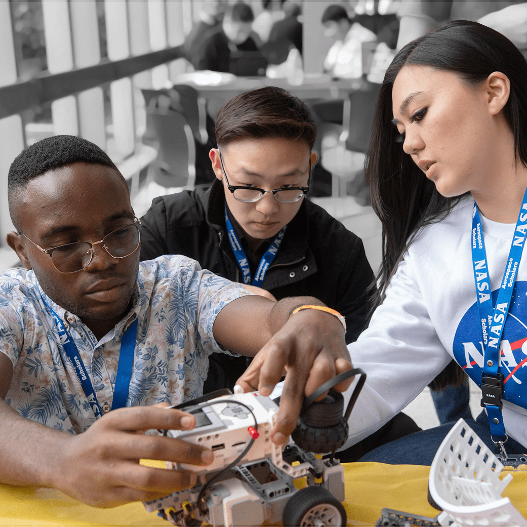 Three students surrounding a small piece of hardware in a lab setting. All students have NASA lanyards on.