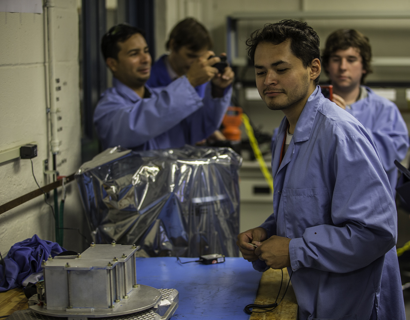 Capitol Technology University students clad in blue engineer scrub coats weigh in their experiment. One student in the foreground weighs a metal box containing the experiment on a blue scale. Three more students are in the background, the student to the far left is taking a photo on a small digital camera. The white walls have multicolored wires plugged into various outlets.