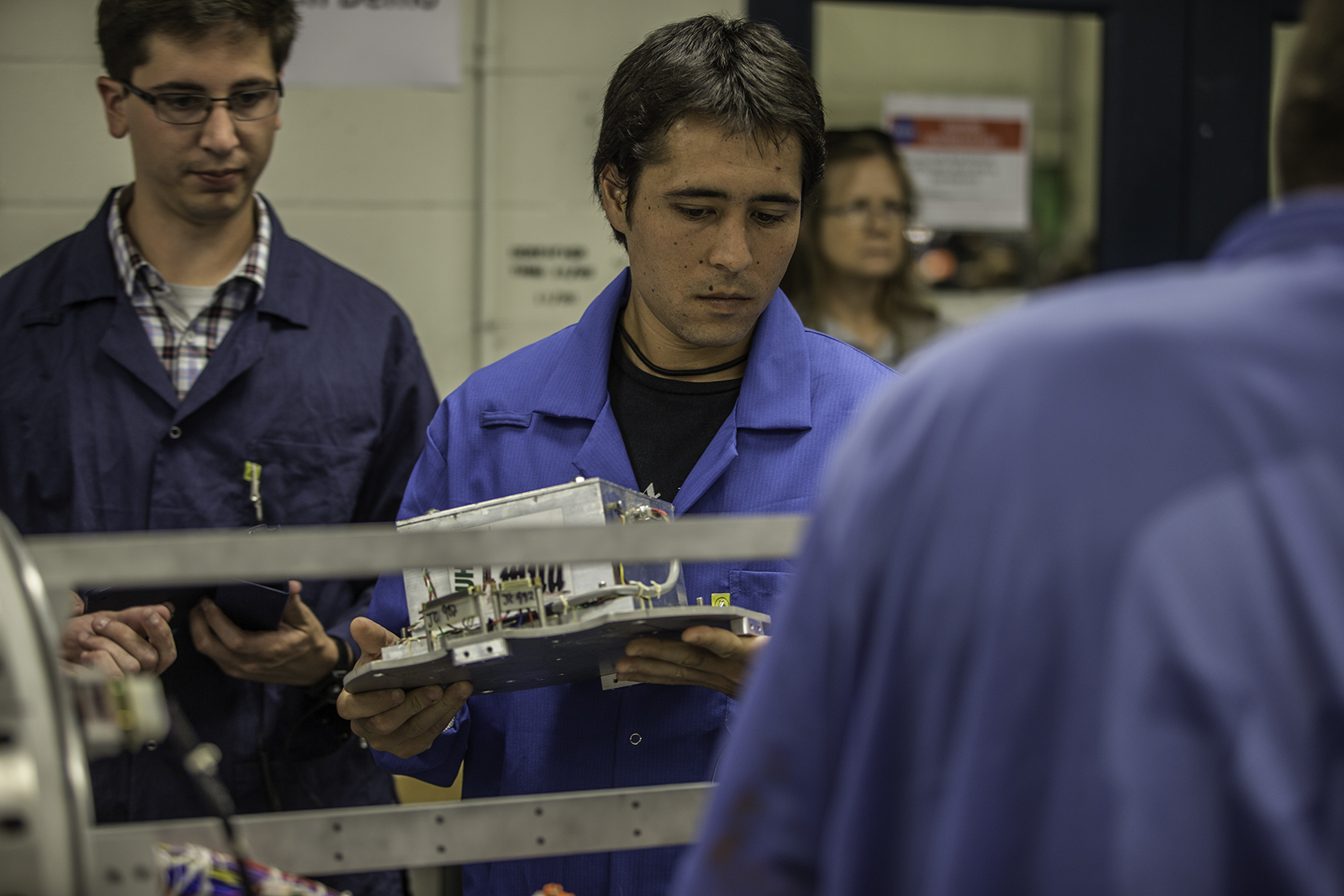 Jesse Austin, program manager Colorado Space ‎Grant Consortium, oversees Marcus Yamaguchi from Kauai Community College, both clad in blue engineer's scrub coats. As he prepares to integrate their experiment, a small white box with multicolored wires, into the payload of a Terrier Improved Malemute sounding rocket, a tubular steel frame.