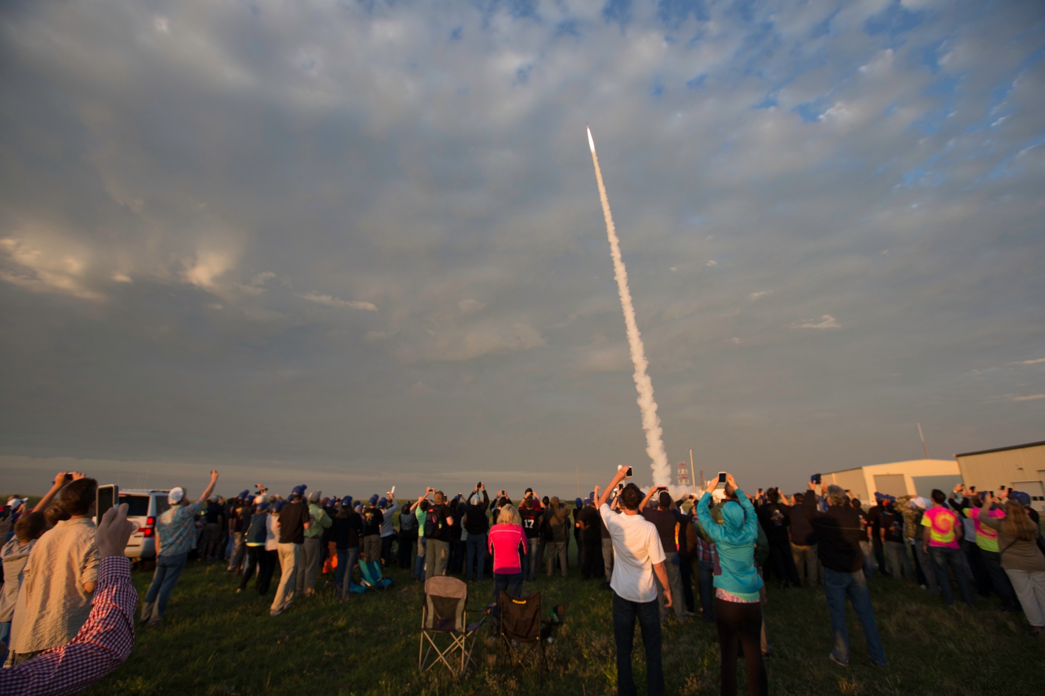 The smoke of a Terrier-improved Malemute rocket is in the top right of the image, streaking upward against a blue sky just beginning to light up at dawn. The bottom of the image is filled edge-to-edge with students excitedly filming the launch on their cell pohones. Two empty camp chairs are in the center of the iamge, abandoned by students too excited to sit.