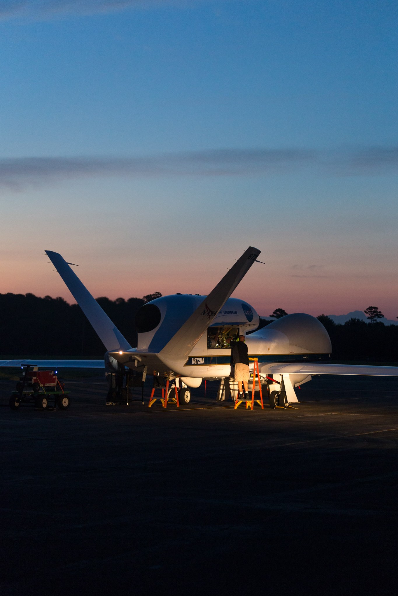 At dusk, crew on orange ladders are gathered around Global Hawk 872 at NASA’s Armstrong Flight Research Center in Edwards, California on a runway, lit up by lamps. The UAV's forked, v-shaped tailfins and wings are visible. In the dark background, brush and bushes are silhouetted against the sunset sky.