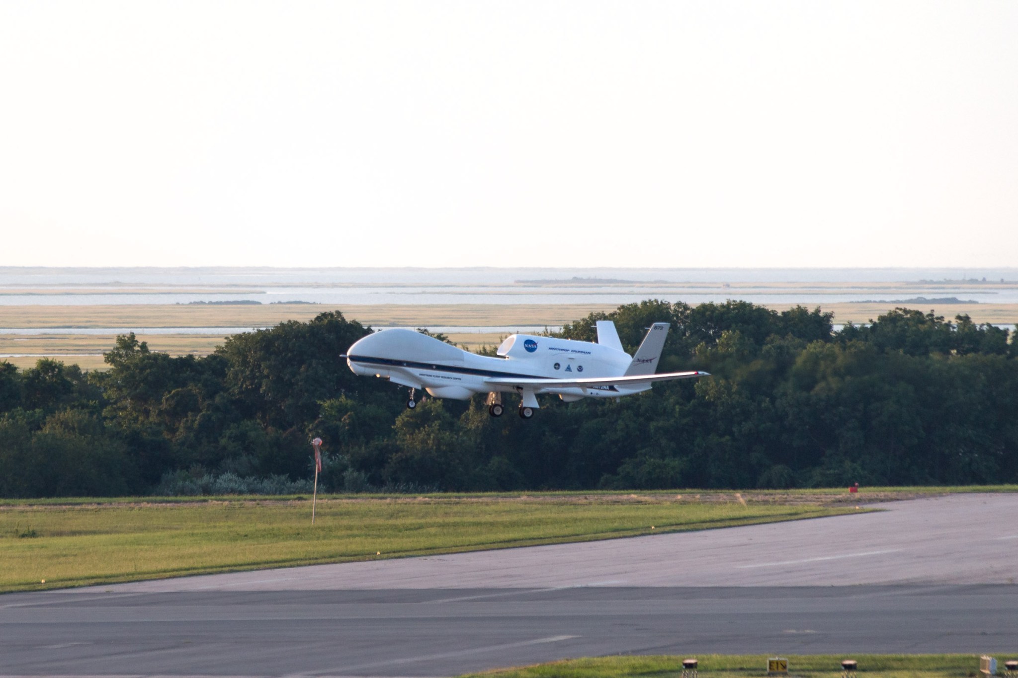 The white, unmanned Global Hawk 872 aircraft takes off from WallopsFlight Facility on a bright clear day with the marshy shoreline in the background. Next to the runway is a pole with a red flag in the middle of the grass. on the airscoop on the top of the aircraft is the nasa meatball. Between the runway area and the shore area is a buffer of thick brush and bushes.