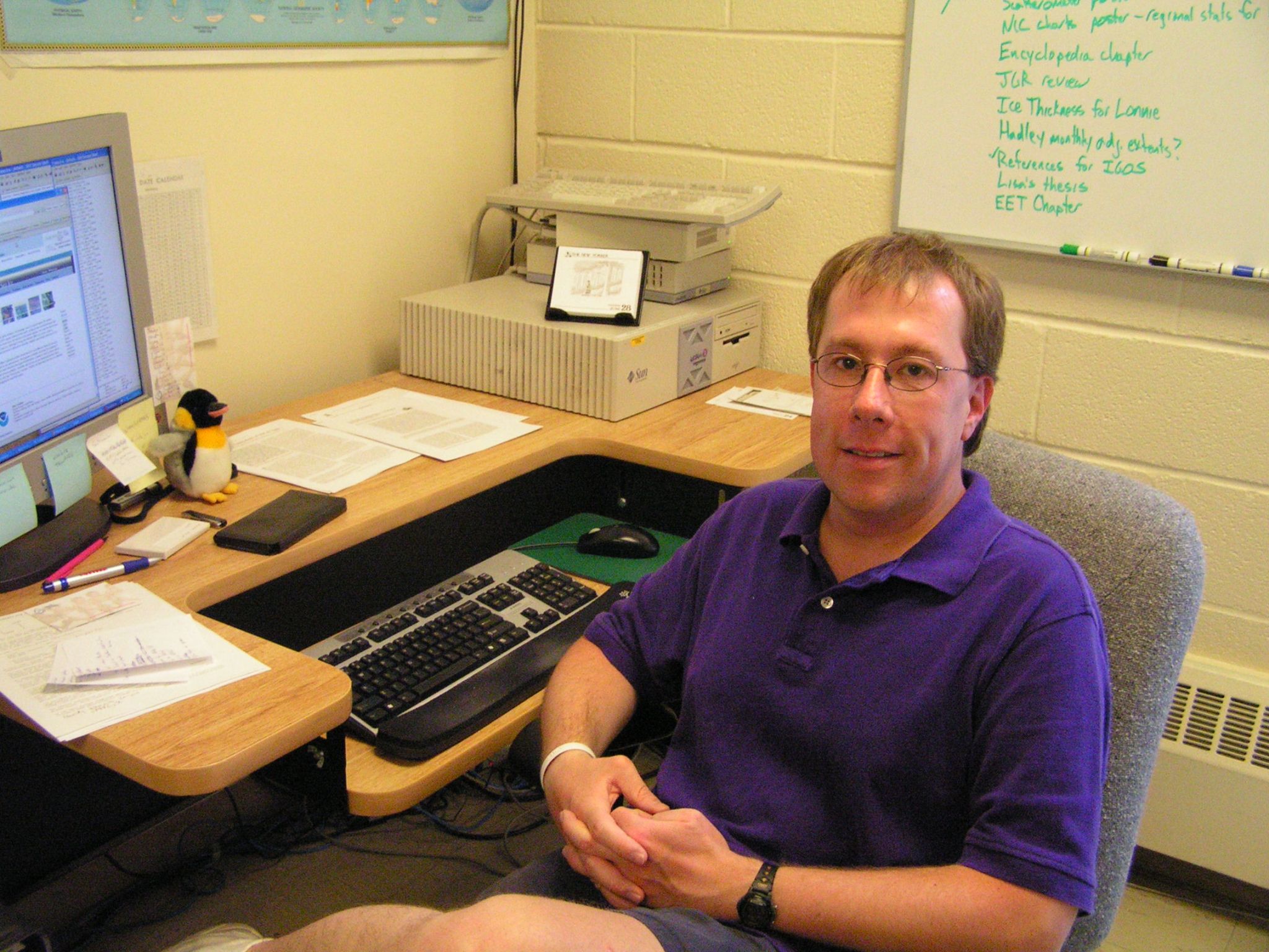 Walt Meier, wearing a purple polo shirt, and sitting at his desk. His hands are folded, and he's smiling at the camera. His desk has a few pieces of paper, a computer monitor, and an older modem.