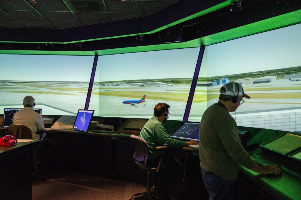 Three tower controllers monitor consoles in FutureFlight Central. The virtual air traffic control tower’s out-the-window views show an aircraft taxiing on the airport’s surface.