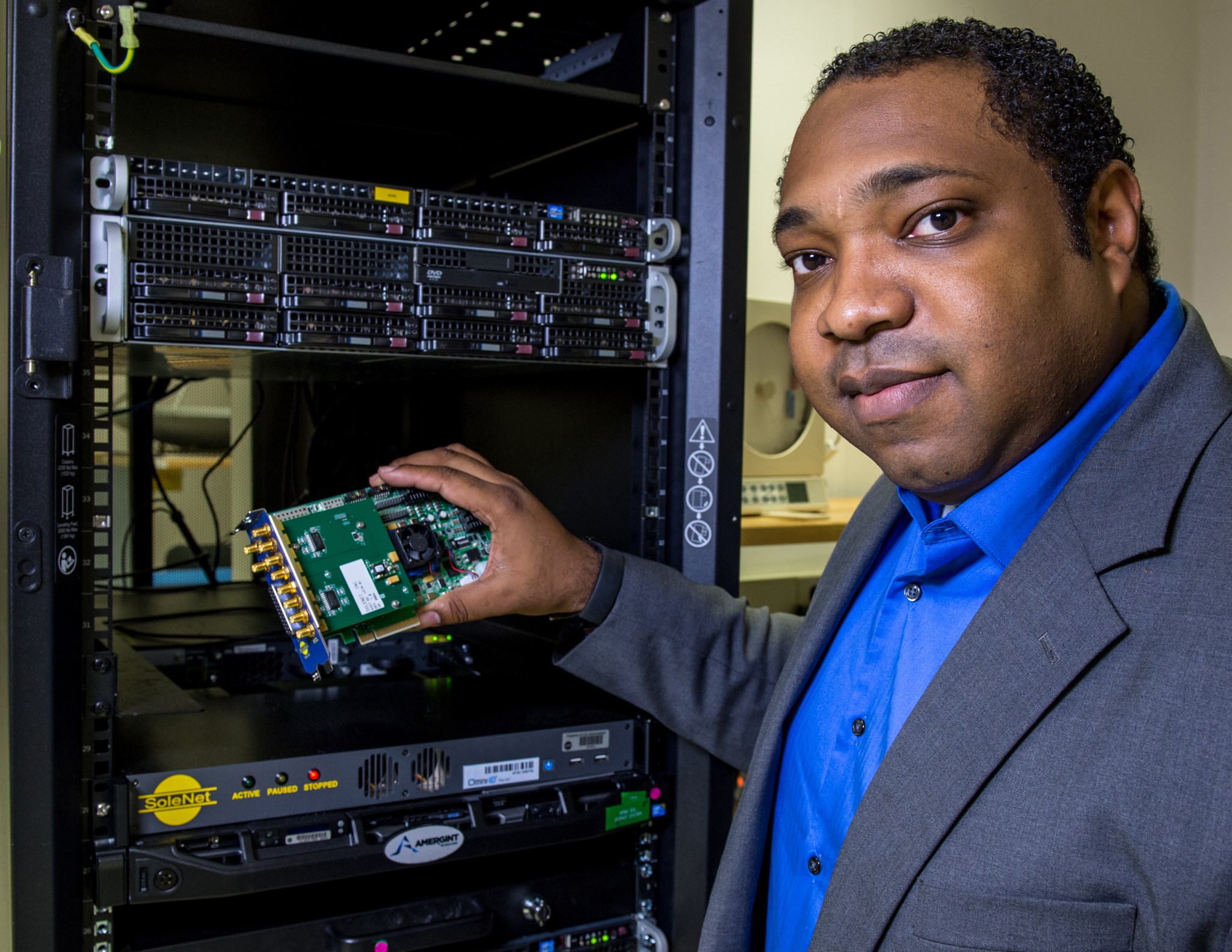 Philip Baldwin holds the custom-designed high-speed interface card. It looks like a green circuit board about the size of a hand. He is turned slightly, looking at the camera over his shoulder.
