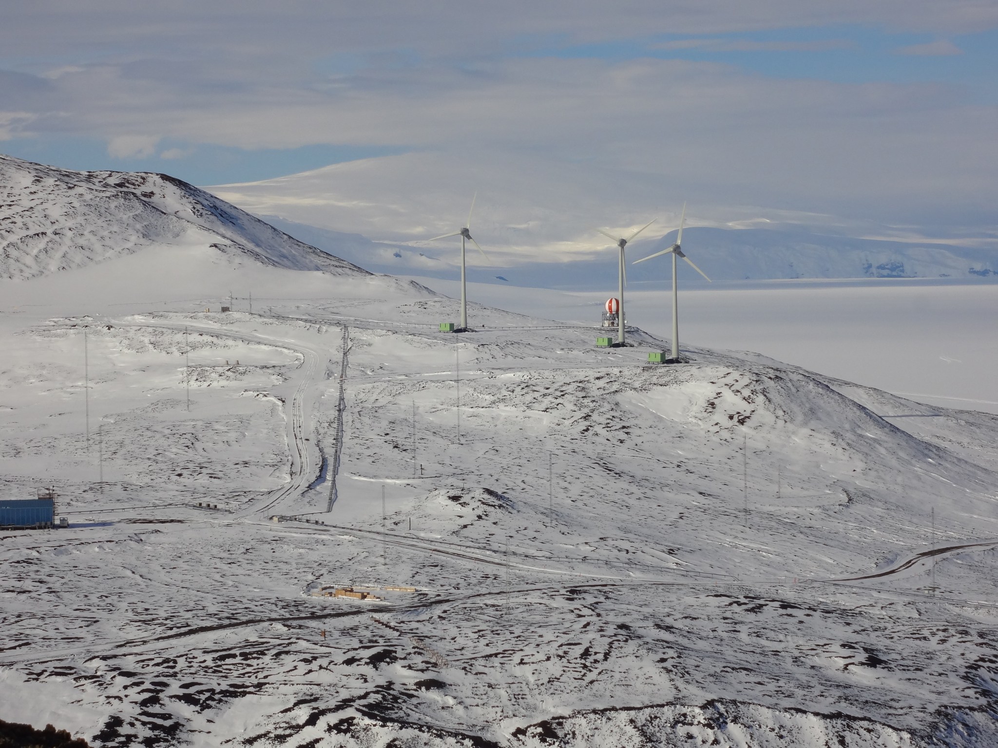 The orange-and-white radome is visible in the distance on a snowy landscape, with low mountains to the left. Three wind turbines sit between the dome and the camera. 