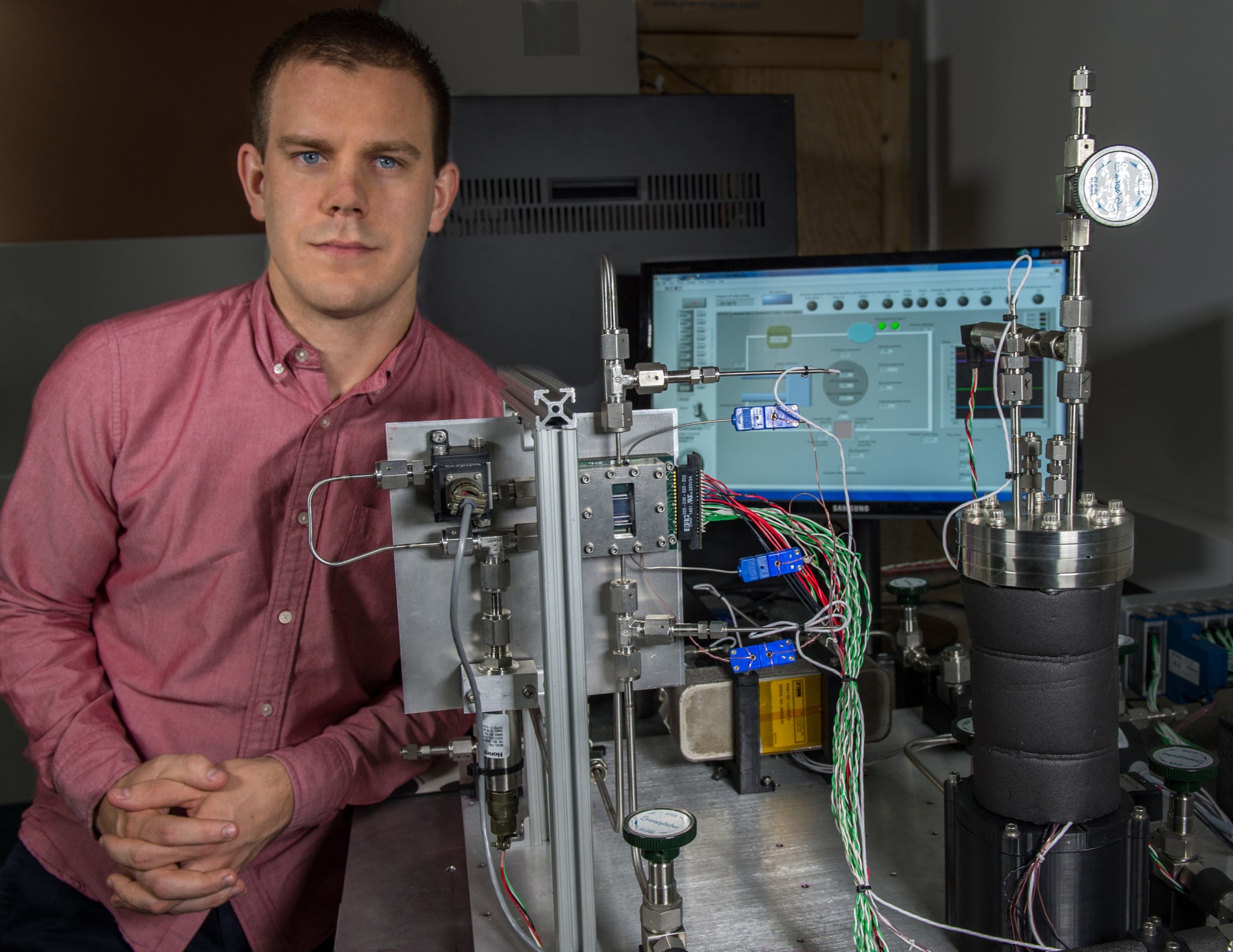 Technologist Franklin Robinson, young man with brown hair wearing a red button down shirt,  poses with a testbed he’s developed to experiment with a new cooling technique for emerging 3-D i