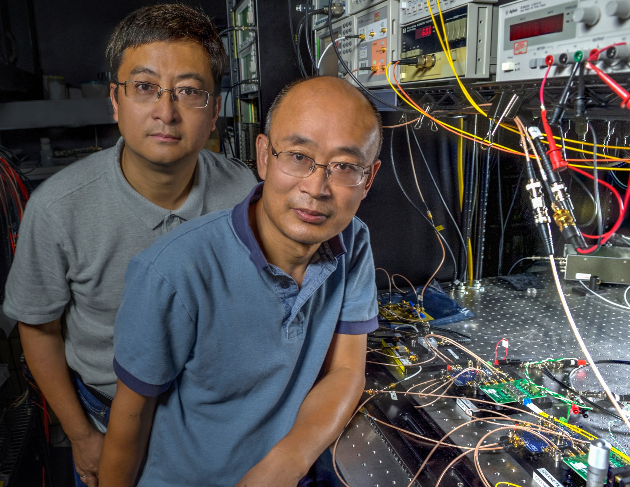 Yang and research associate, Wei Lu both wearing polo shirts and glasses stand with many wires and electrical components
