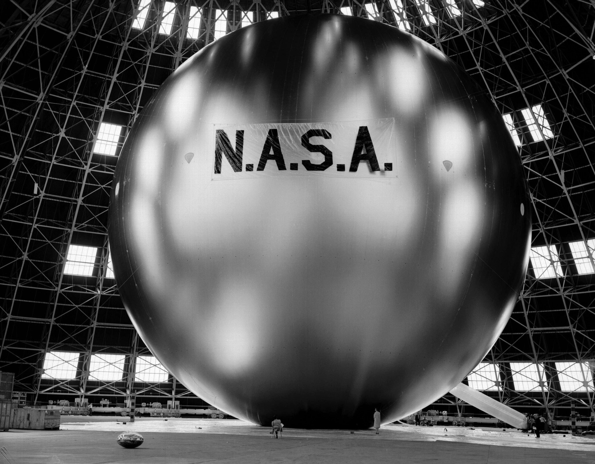 A black-and-white photo shows a very large, silver, spherical communication satellite with N.A.S.A in black letters across the center. The satellite is inside of a large hanger with several windows. Two people can be seen in front of the satellite, appearing very small in comparison.