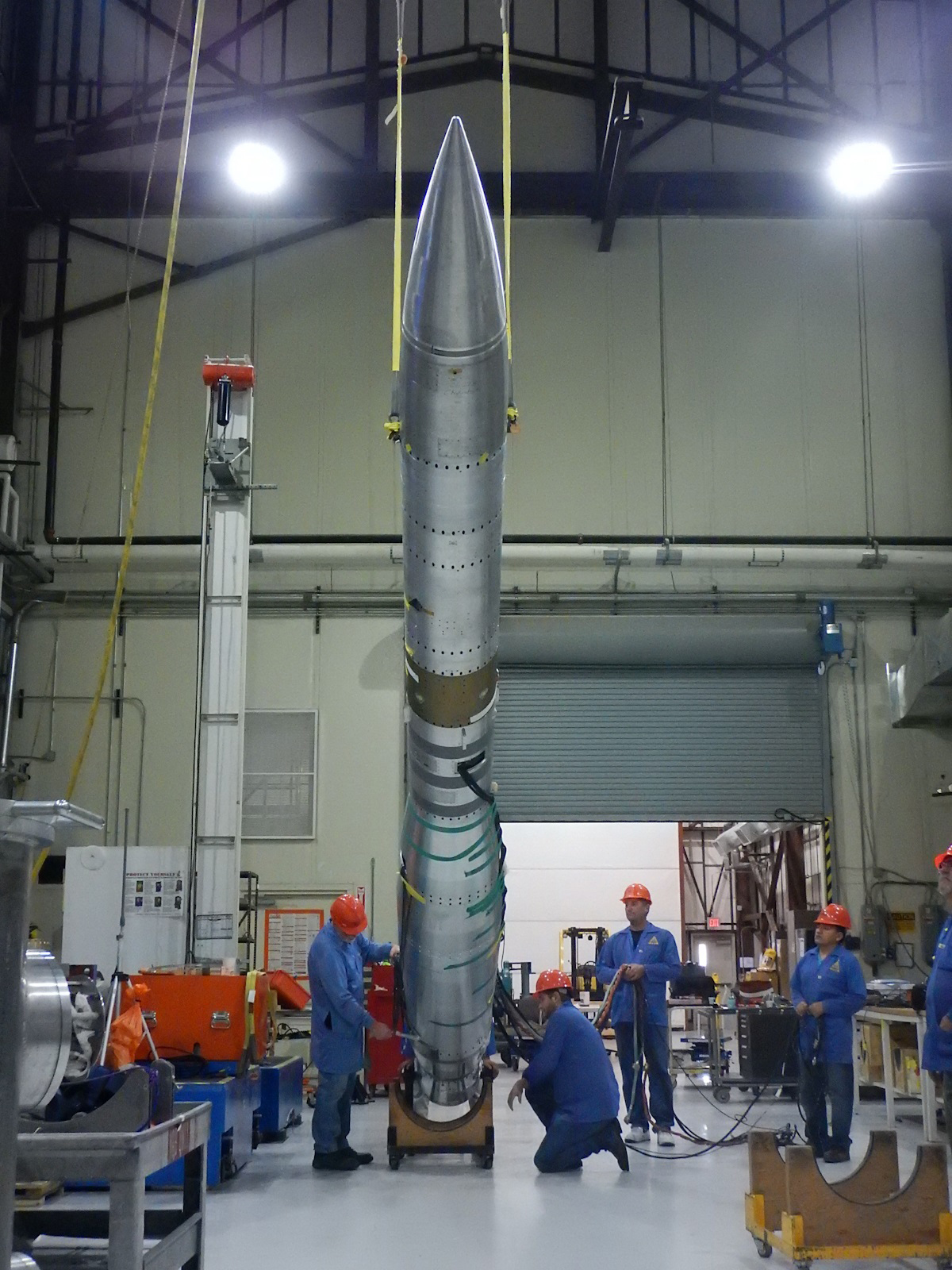 The Planet Imaging Coronagraphic Technology Using a Reconfigurable Experimental Base (PICTURE-B) is integrated into a Black Brant IX rocket inside the integration facility at White Sands Missile Range. The Picture-B payload is inside a large, shiny, steel rocket that takes up the center of the image, suspended by yellow safety straps, five workers in blue engineer's scrubs and orange hard hats are cluster around the bottom, one on the left and four on the right. the grey shutter door in the bottom right quarter of the background is half-open. the workshop area is filled with electrical equipment.