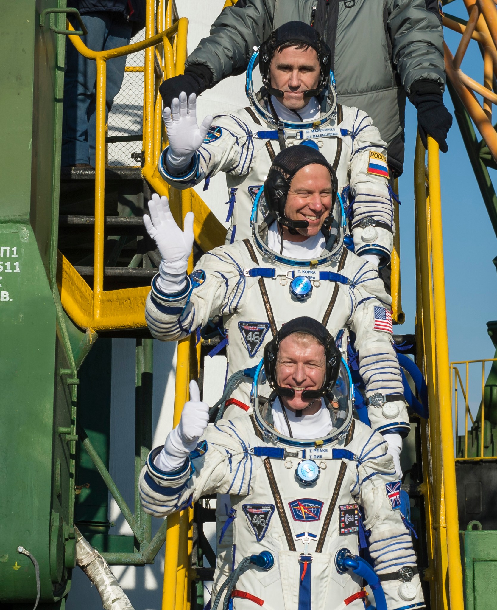 Three men in Sokol launch suits wave while standing on stairs leading up to Soyuz capsule