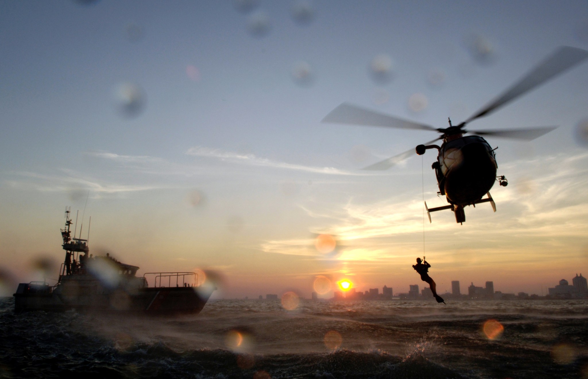 A helicopter flies low above the ocean during sunrise. A rescue diver is suspended from the helicopter, and nearby, a boat floats on the surface of the water. The skyline of a city can be seen in the far distance against a sky of blue and gold hues. Water droplets can be seen on the lens of the camera capturing the image.