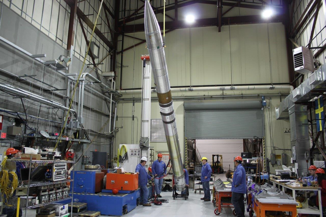 Four men stand around a diagonally-suspended body of a Black Brant IX suborbital sounding rocket that carries the Colorado High-resolution Echelle Stellar Spectrograph, CHESS-2, in the rocket integration area at White Sands Missile Range. The men are wearing hard hats colored (from left to right) white, red, yellow and red and blue engineering scrub coats. Two other people are seated to the very furthest right of the image wearing red hard hats. The workshop is full of orange and blue workbenches, steel shelves full of electrical equipment and other crates and tools.
