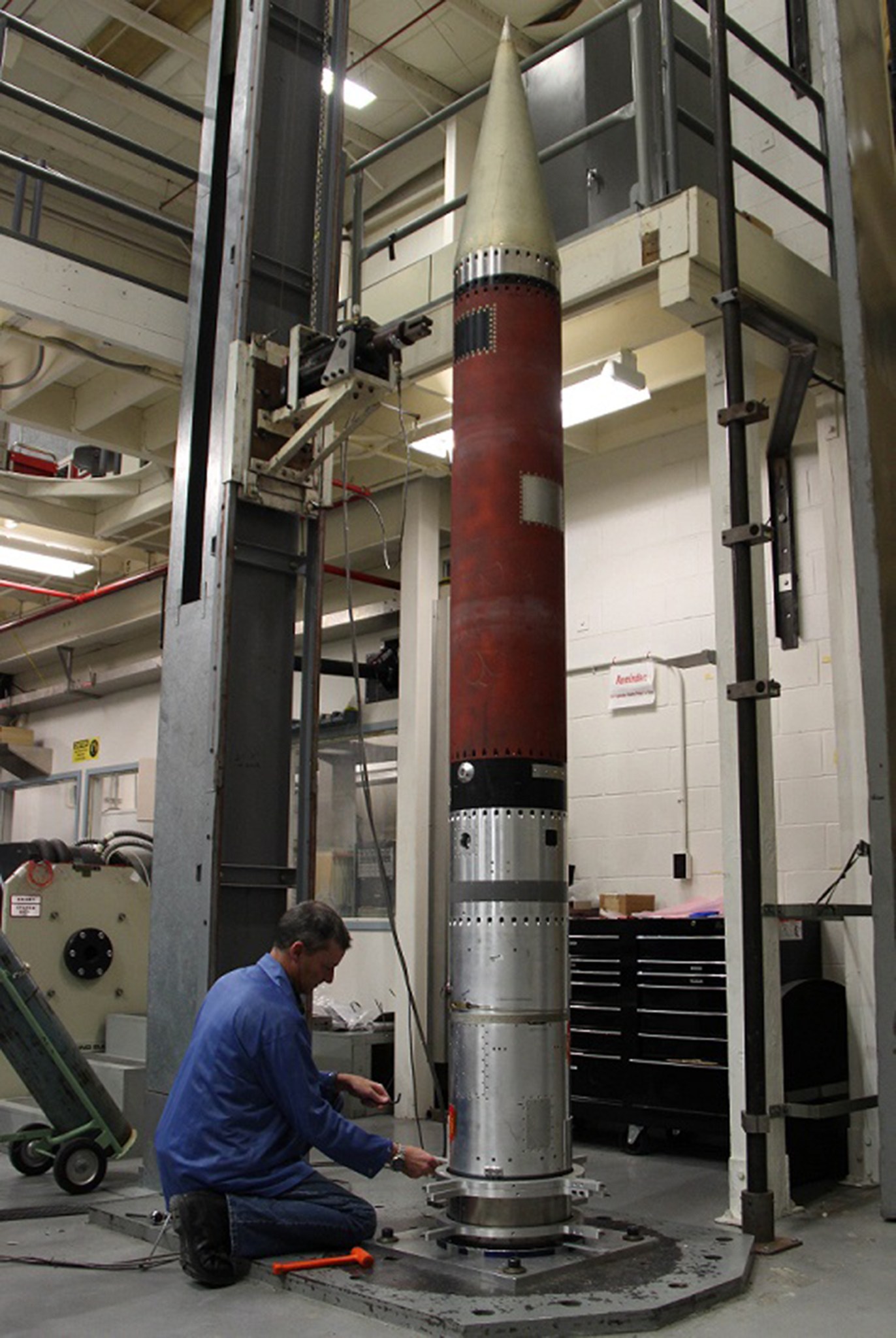 A person is sitting at the base of a rocket payload that is sitting vertical on a platform.