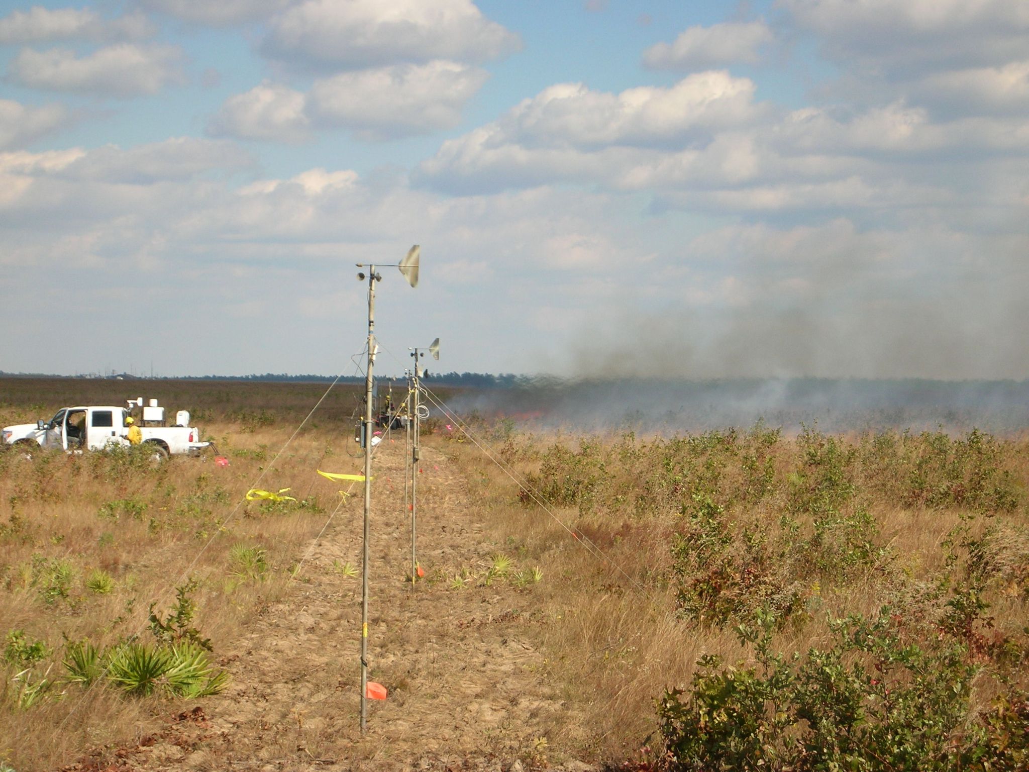 Fire being set by a fire boss riding on an off-road vehicle fairly visible in line with and beyond the wind vanes.