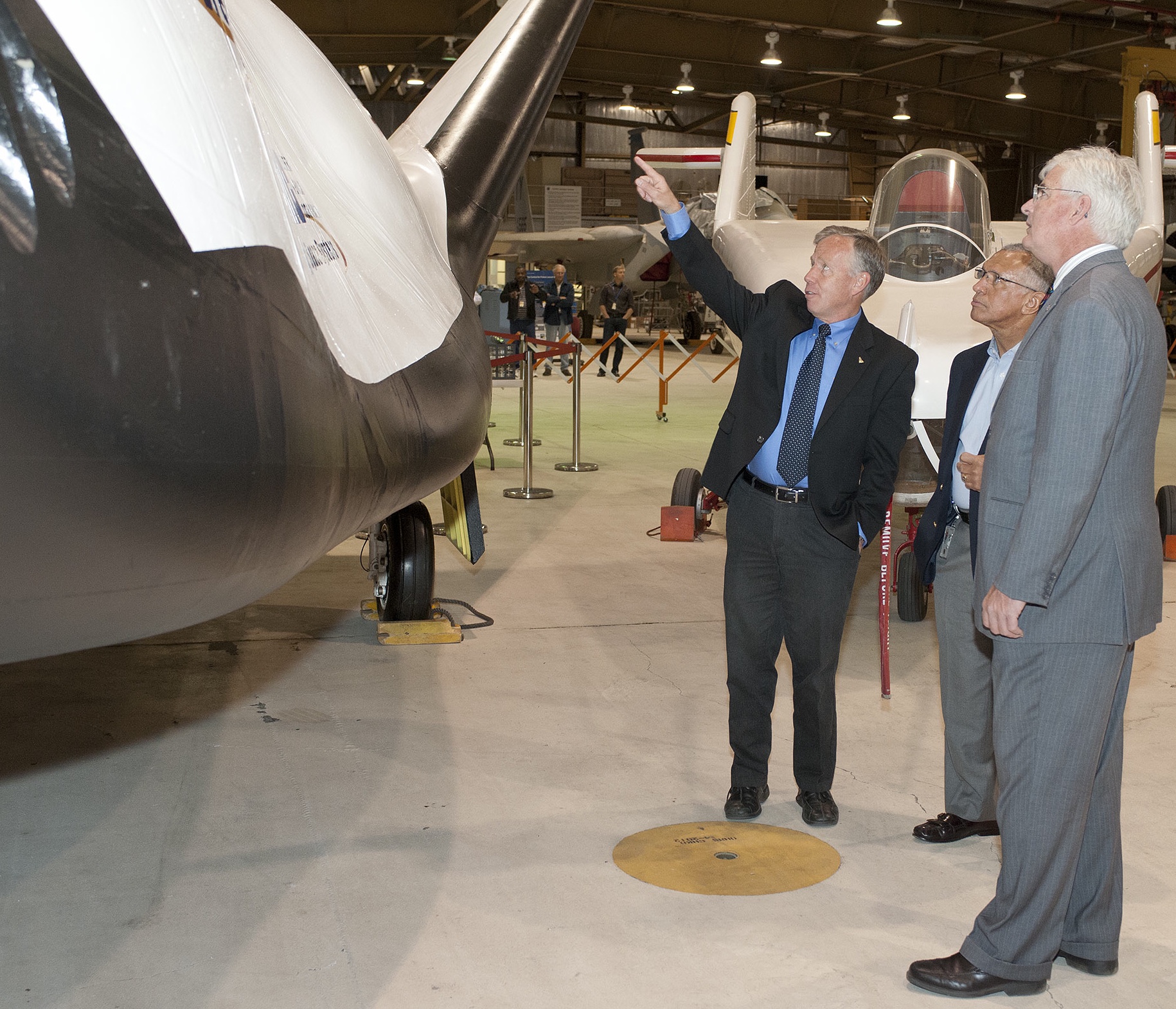Astronaut Steve Lindsey points out features of the firm's prototype Dream Chaser flight test vehicle to then NASA Administrator Charlie Bolden and Patrick Stoliker. Behind them adjacent to the Dream Chaser is the original M2-F1 lifting body aircraft.