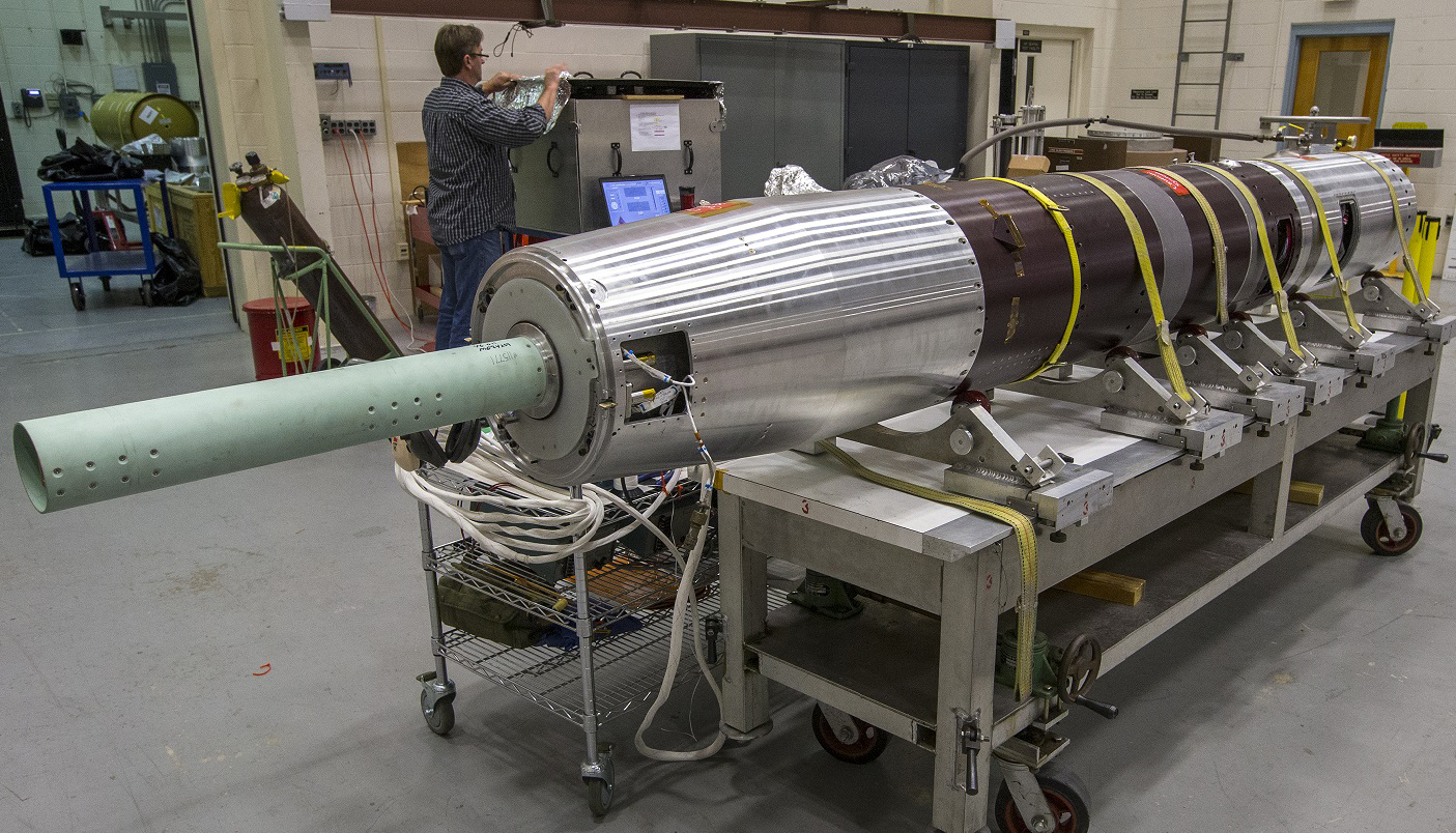 A partially assembled sounding rocket payload containing the Multiple User Suborbital Instrument Carrier (MUSIC) sits securely on a workbench in the sounding rocket manufacturing are at Wallops Flight Facility. A man in a navy-and-white striped shirt is in the background, opening a container. Behind the rocket and the man is a white-walled workshop full of carts, security straps, tanks of liquids and gasses and other machine equipment.