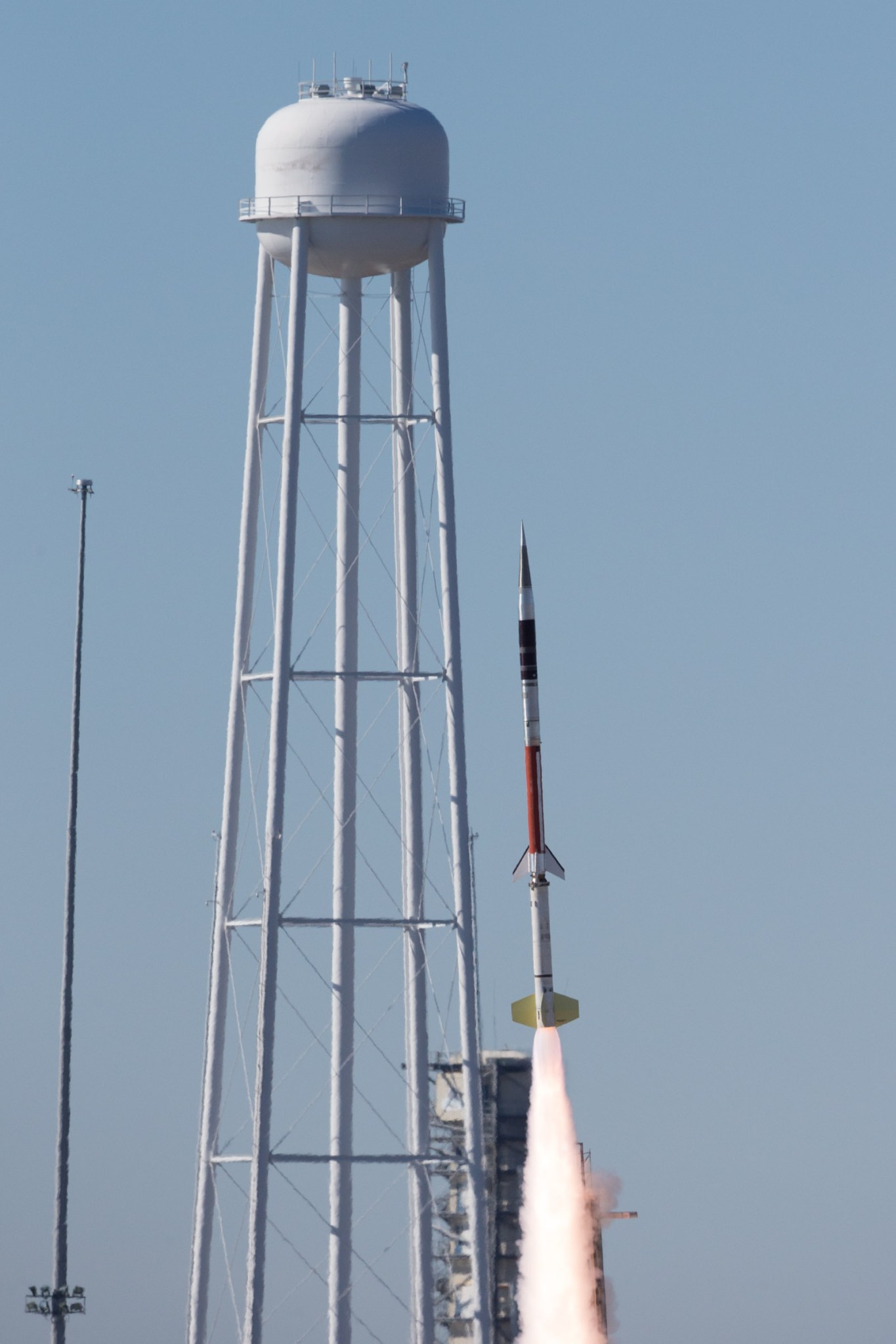 A Terrier-Improved Malemute suborbital sounding rocket launches with the MUSIC mission from the rocket launching facility on the south end of Wallops Island at Wallops Flight Facility. In the background against a blue sky is a water tower that appears to be three or four times the height of the rocket. In the left of the image is a vertical grey pole.