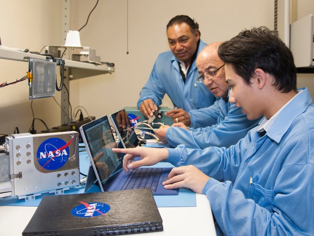 Three men in blue lab coats sitting in front of laptops