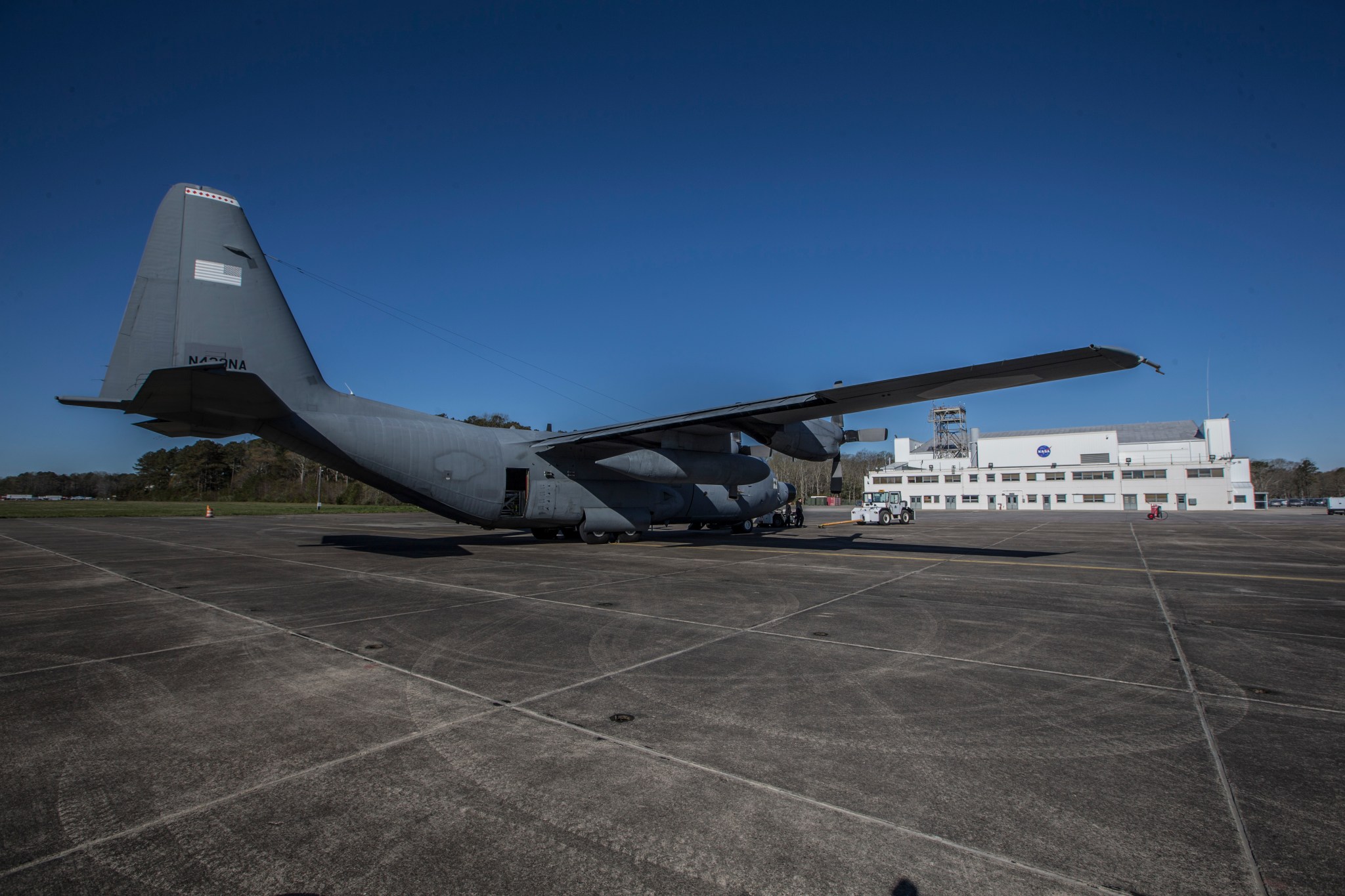 A C-130 aircraft sits on a runway at Wallops Flight facility with the nose pointing towards the rear right of the image. The tailfin, closer and to the left in the image has a faded American flag. In the background of the photo in front of a clear blue sky is a large white building with the NASA meatball logo painted on the exterior on the upper floor.