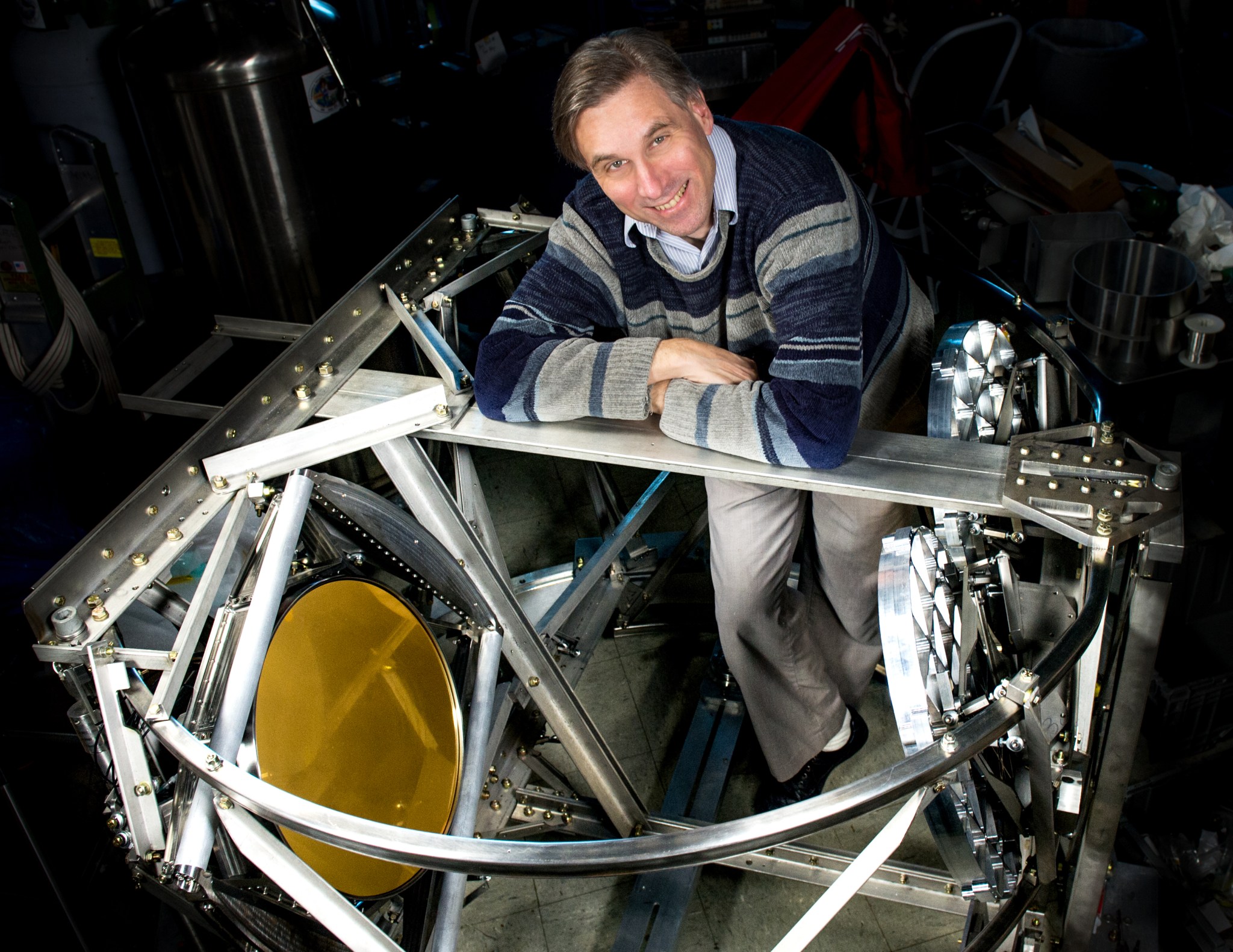 Al Kogut leans on a rounded metal frame with a gold lens on the side. He is smiling up at the camera.