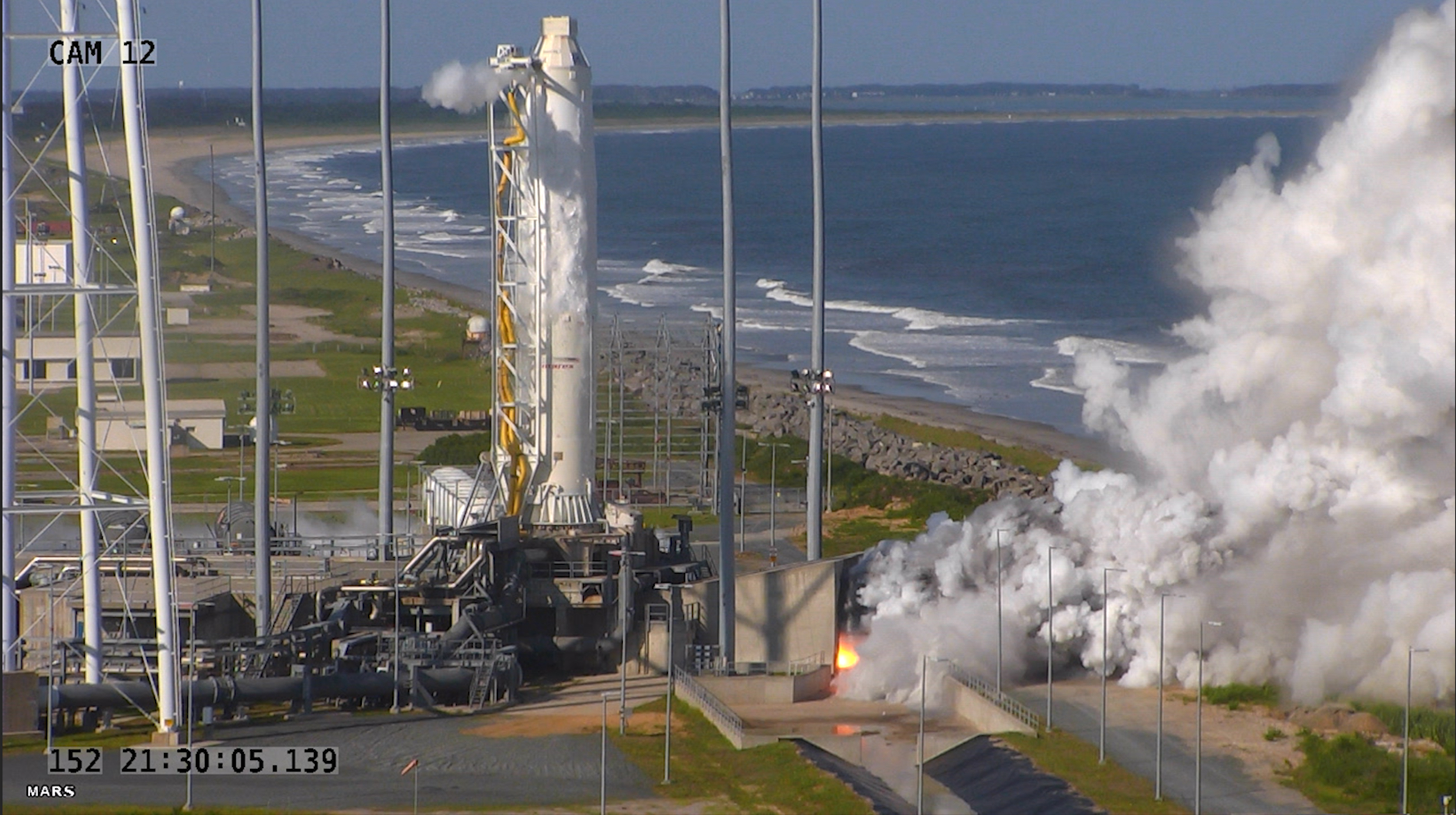 A screenshot of a video feed showing the first stage of a rocket attached to a the pad, with a flaming shooting out of the side with a large plume of smoke.